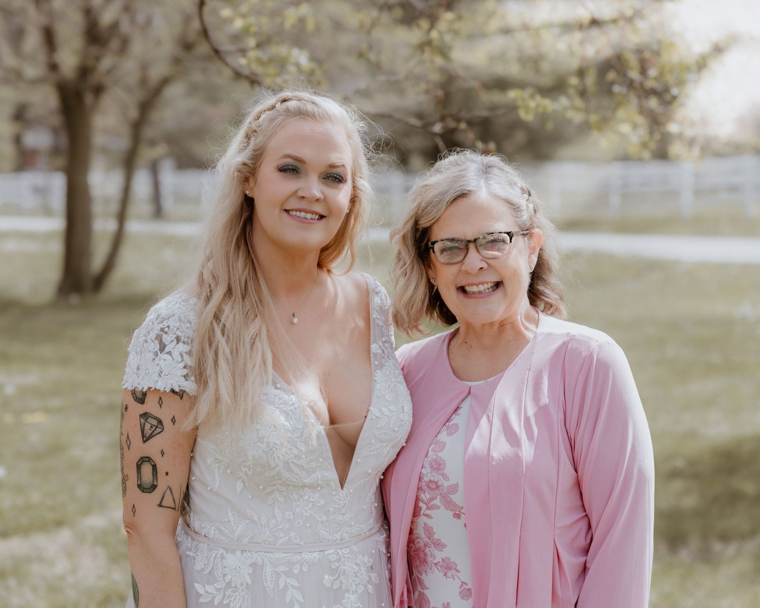 Bride in white lace gown poses with older woman in pink cardigan outdoors at spring wedding.
