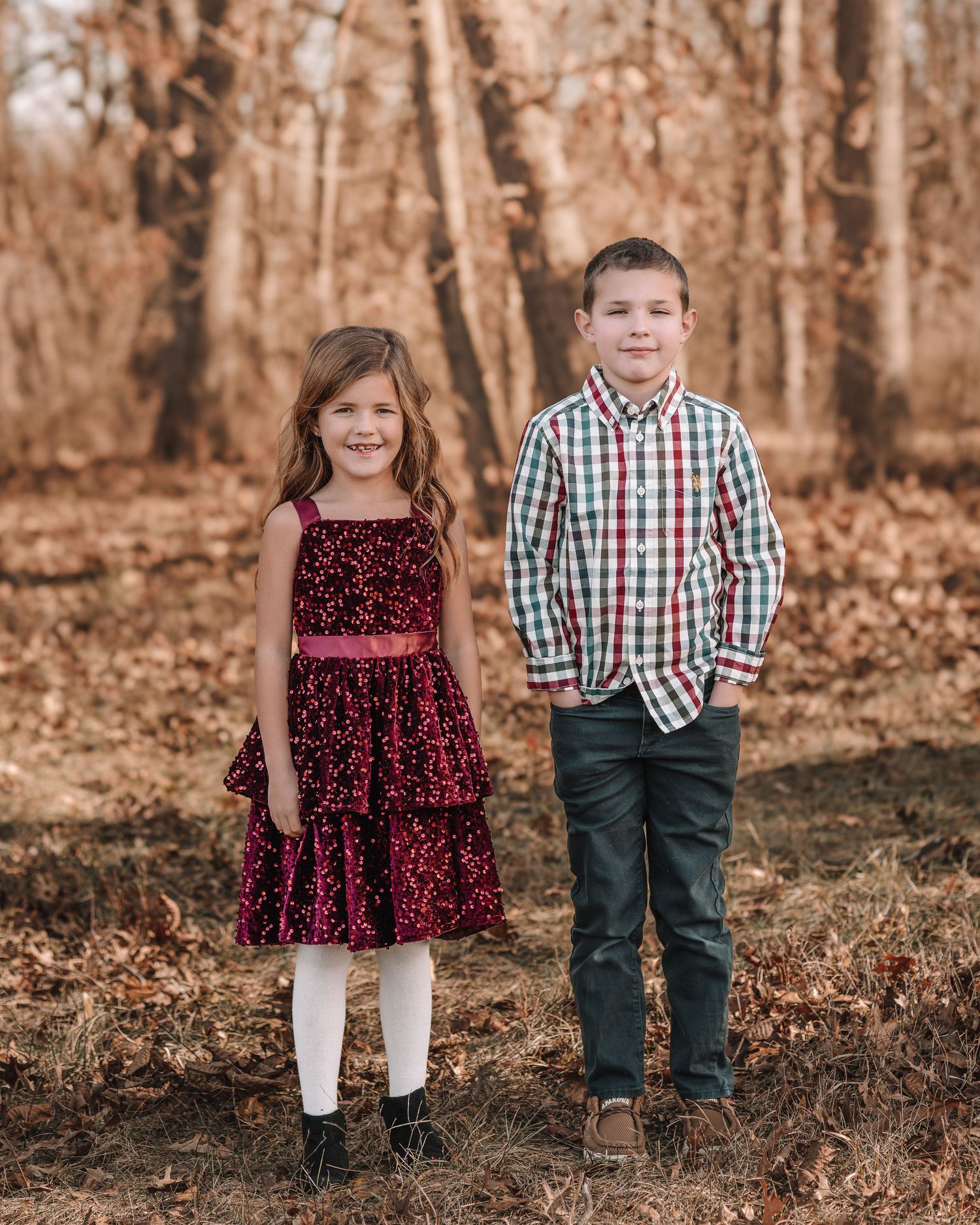 Two children in coordinating holiday outfits pose together in a wooded autumn setting.