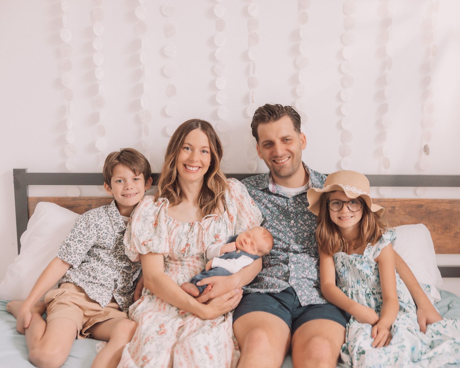 Happy family of five sitting on a bed, parents holding newborn baby flanked by two smiling children.