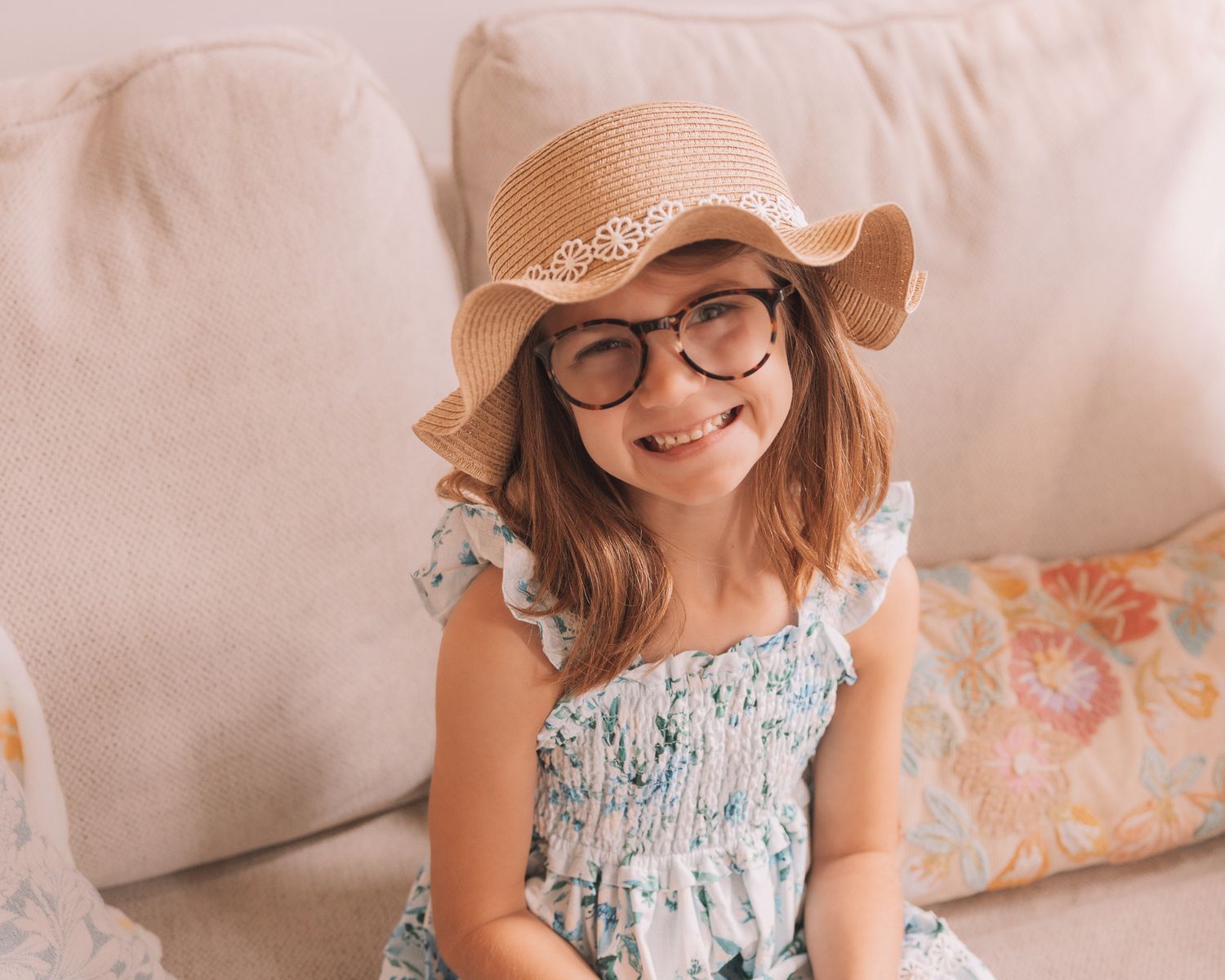 Adorable young girl wearing glasses and a straw hat smiles on a couch.
