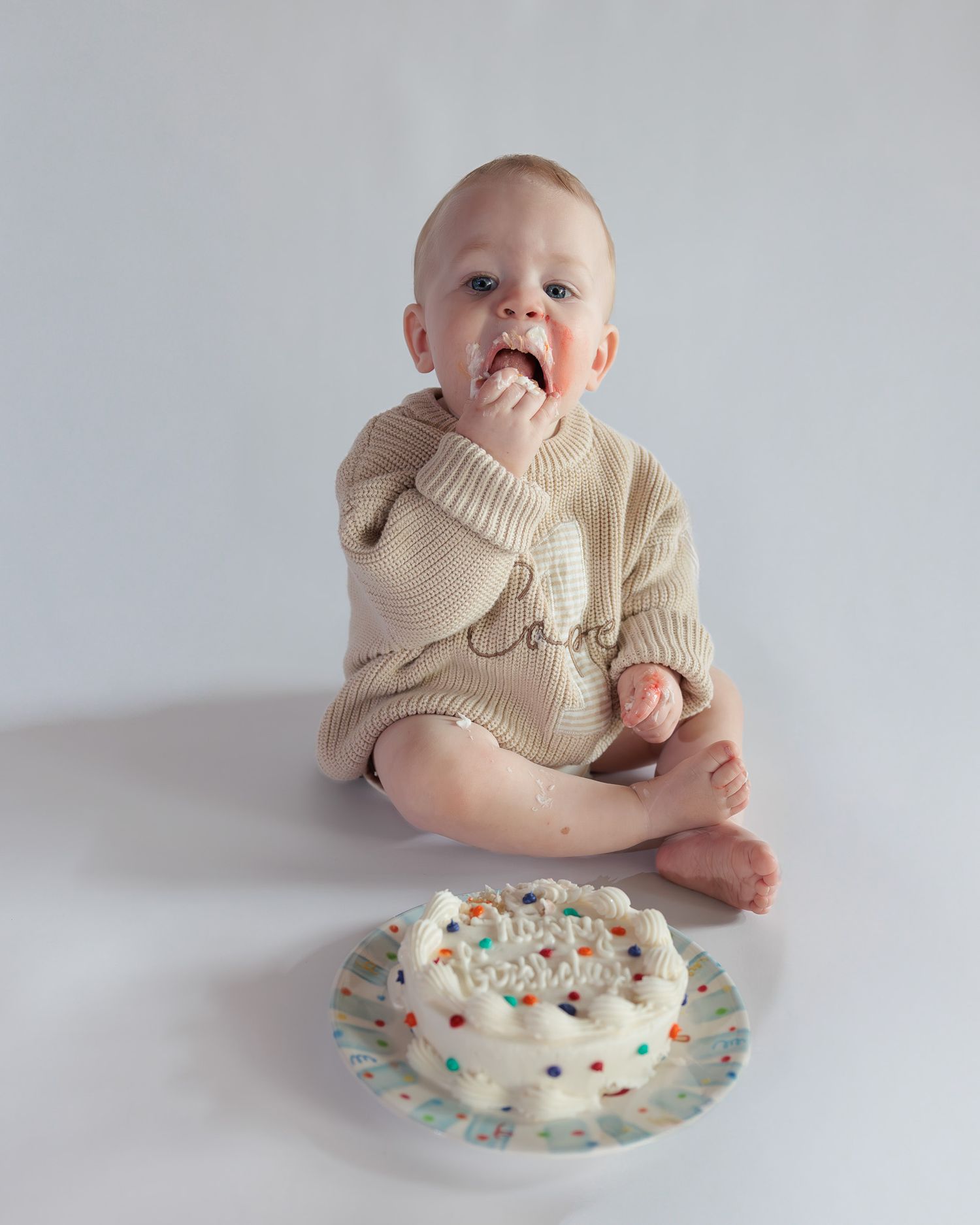 Baby in beige knit sweater eating cake during first birthday smash cake photoshoot.