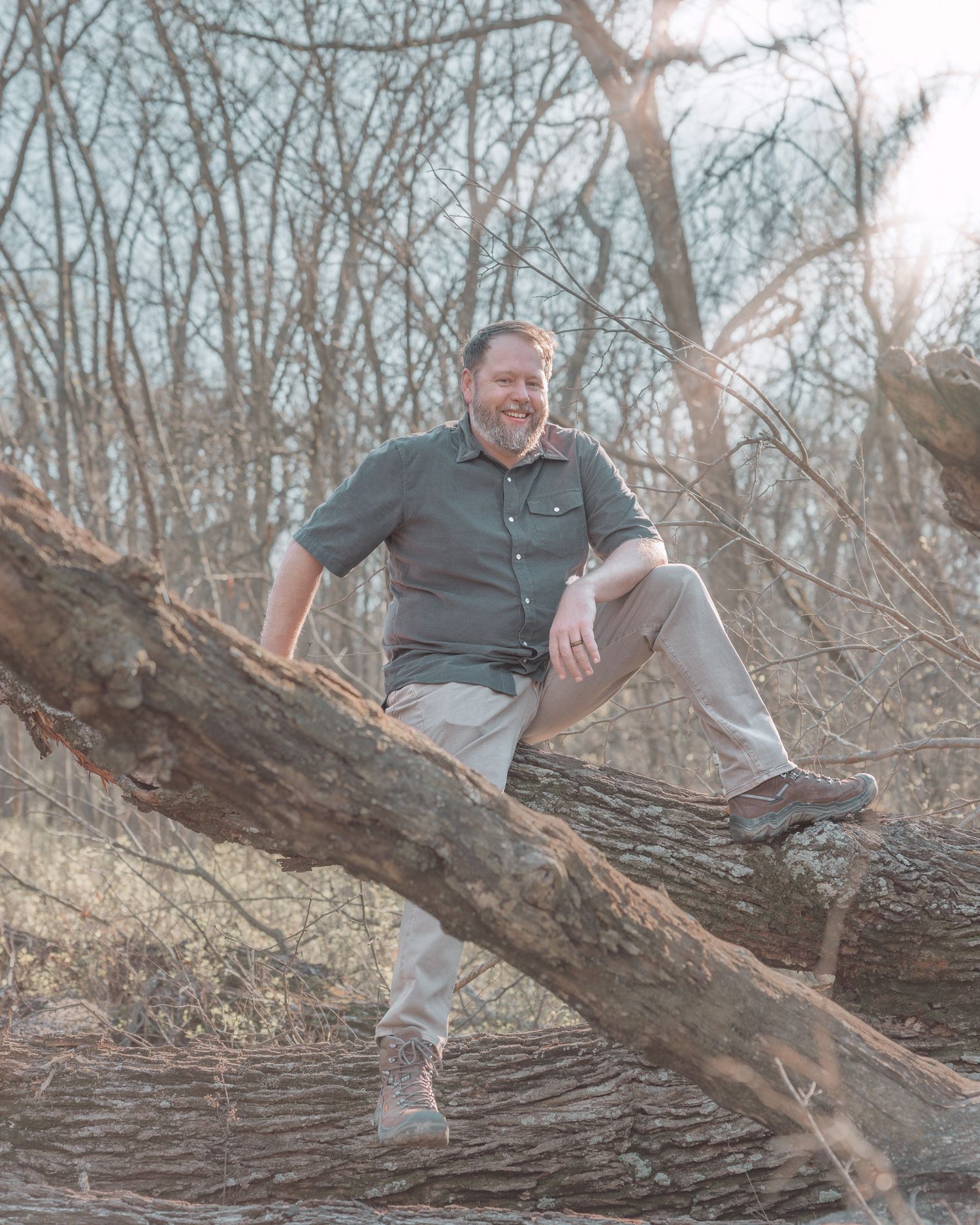 Smiling bearded man sitting relaxed on a fallen tree in a wooded area.