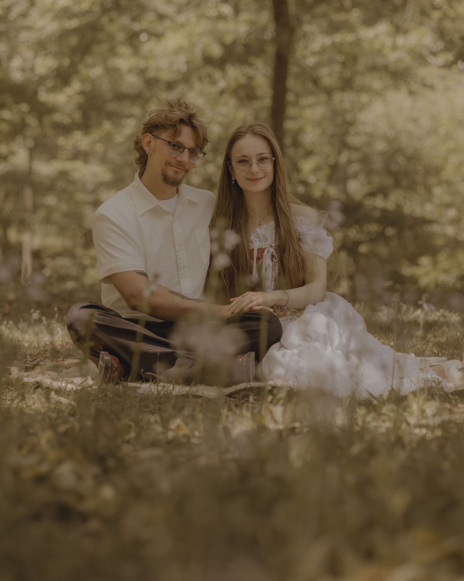 A couple sits together on grass in a sunlit forest, the woman in a white dress and the man in casual attire, both wearing glasses.