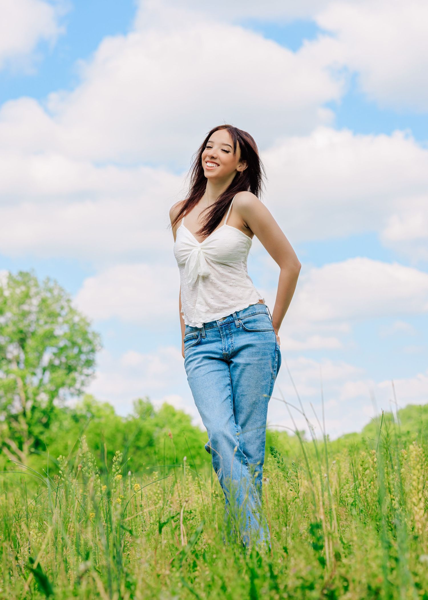 Young woman in white top and jeans smiling in a green meadow under a blue sky with clouds.