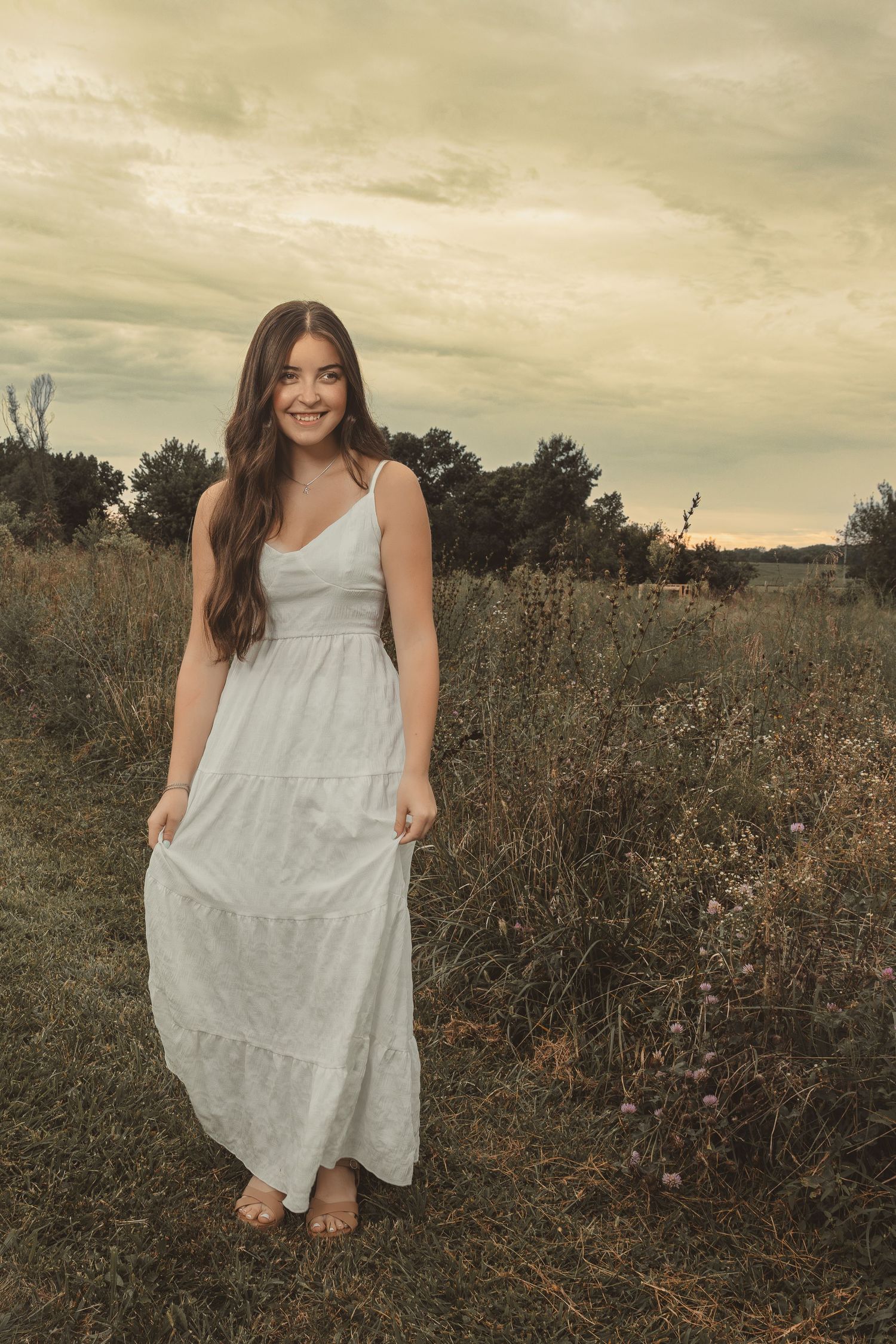 Young woman in white maxi dress smiling in a rustic field with overcast sky and trees in background.