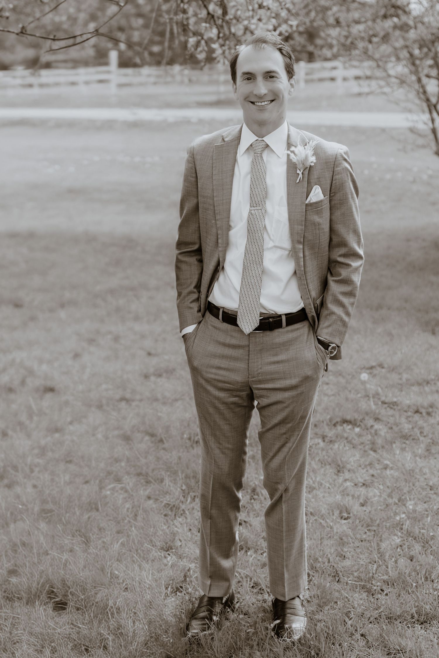 Smiling groom in a tailored suit with striped tie and boutonniere stands outdoors on grass in a sepia-toned wedding photo.