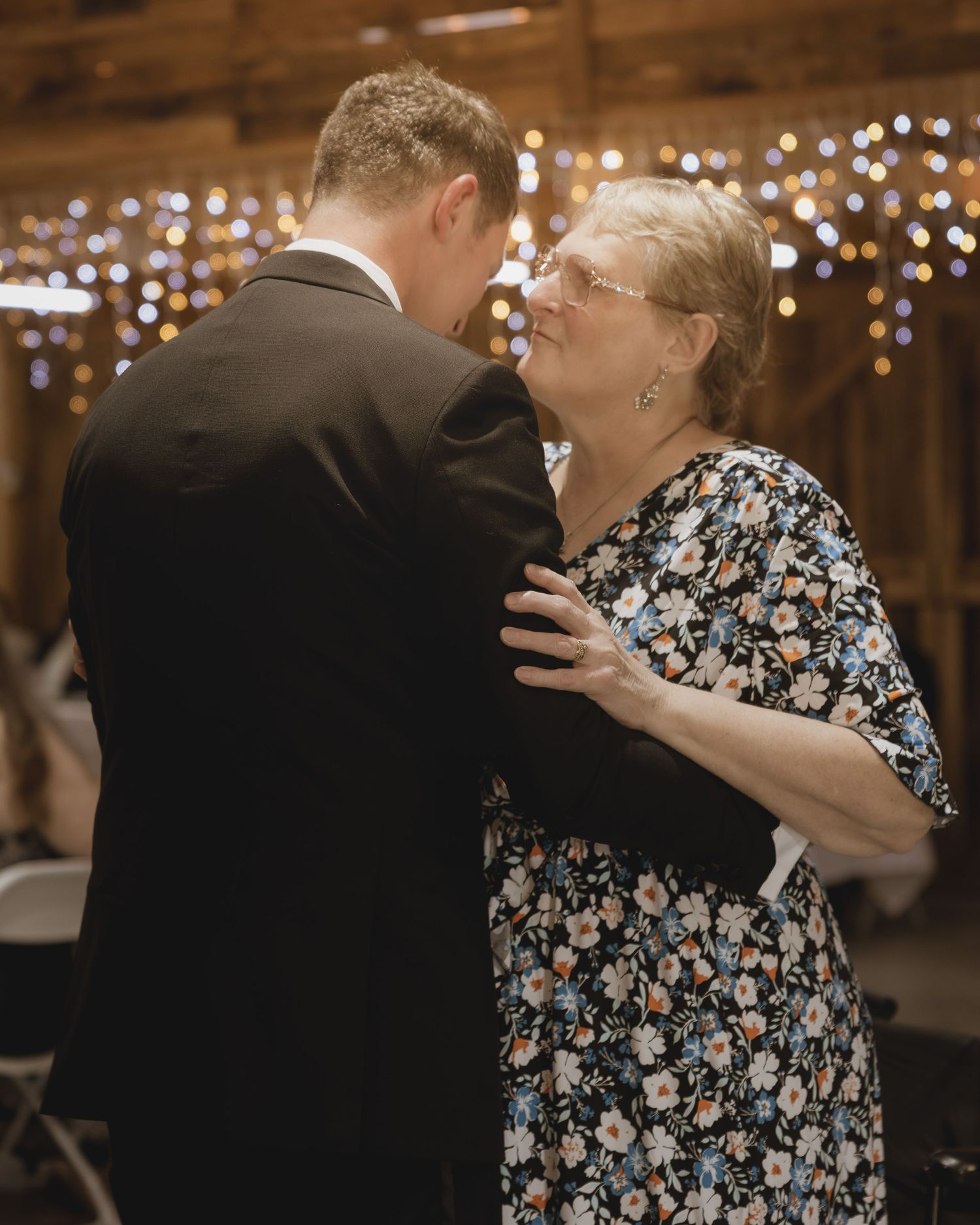Elderly woman and man in suit share a slow dance at a rustic barn wedding reception with twinkling lights.