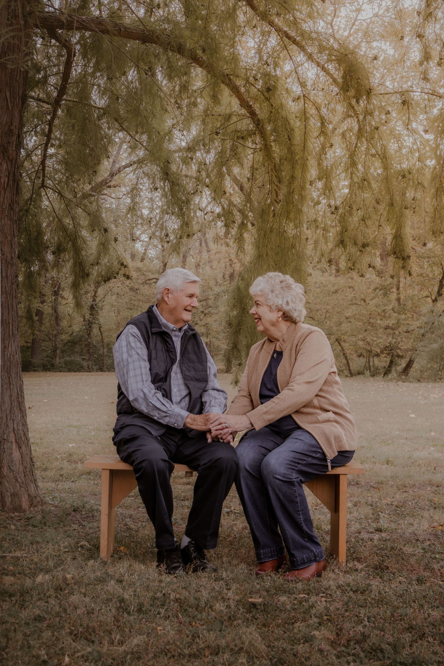 Elderly couple sitting together on wooden bench in park holding hands during autumn afternoon.