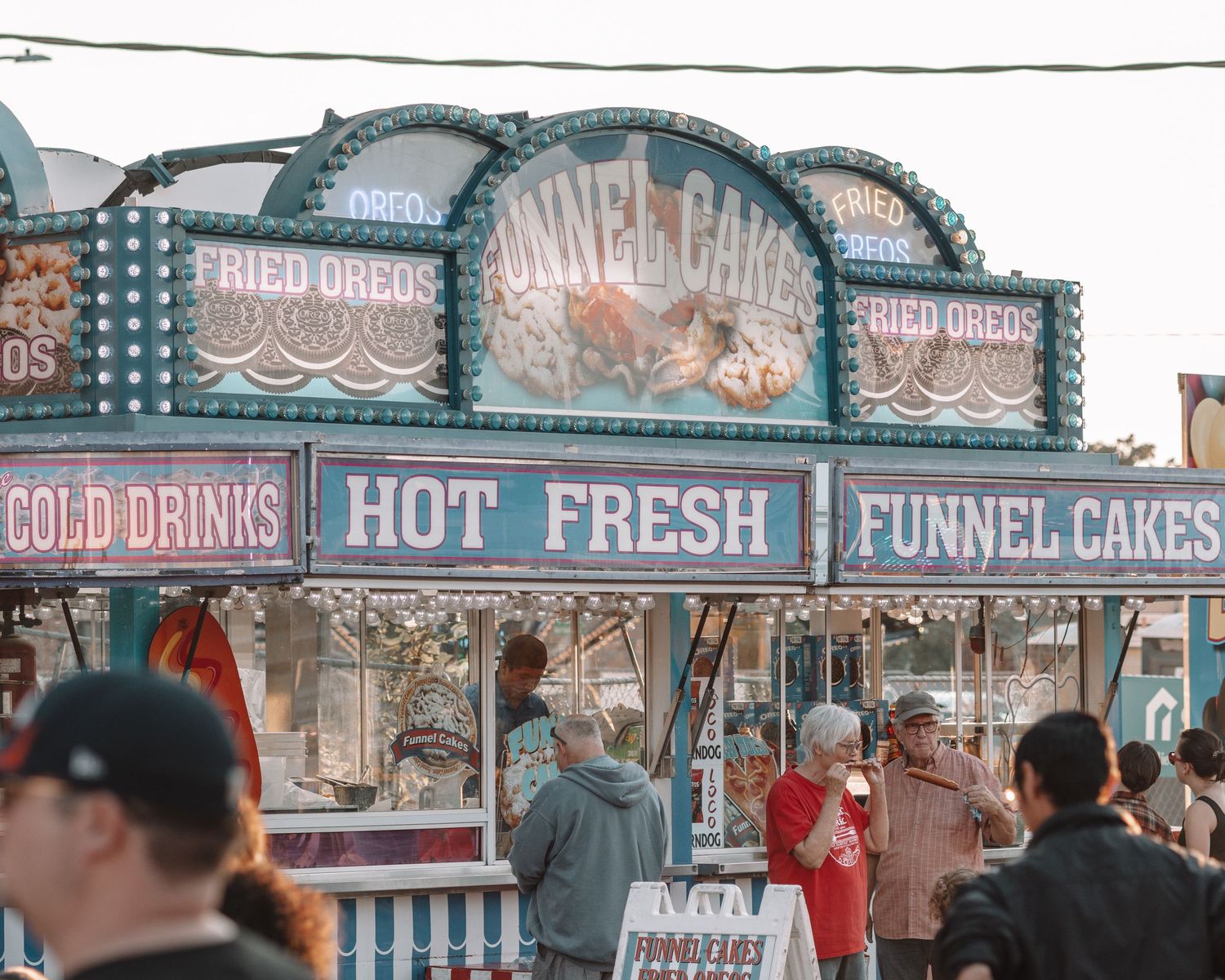 Vintage carnival food stand with signs advertising funnel cakes, cold drinks, and hot fresh treats.