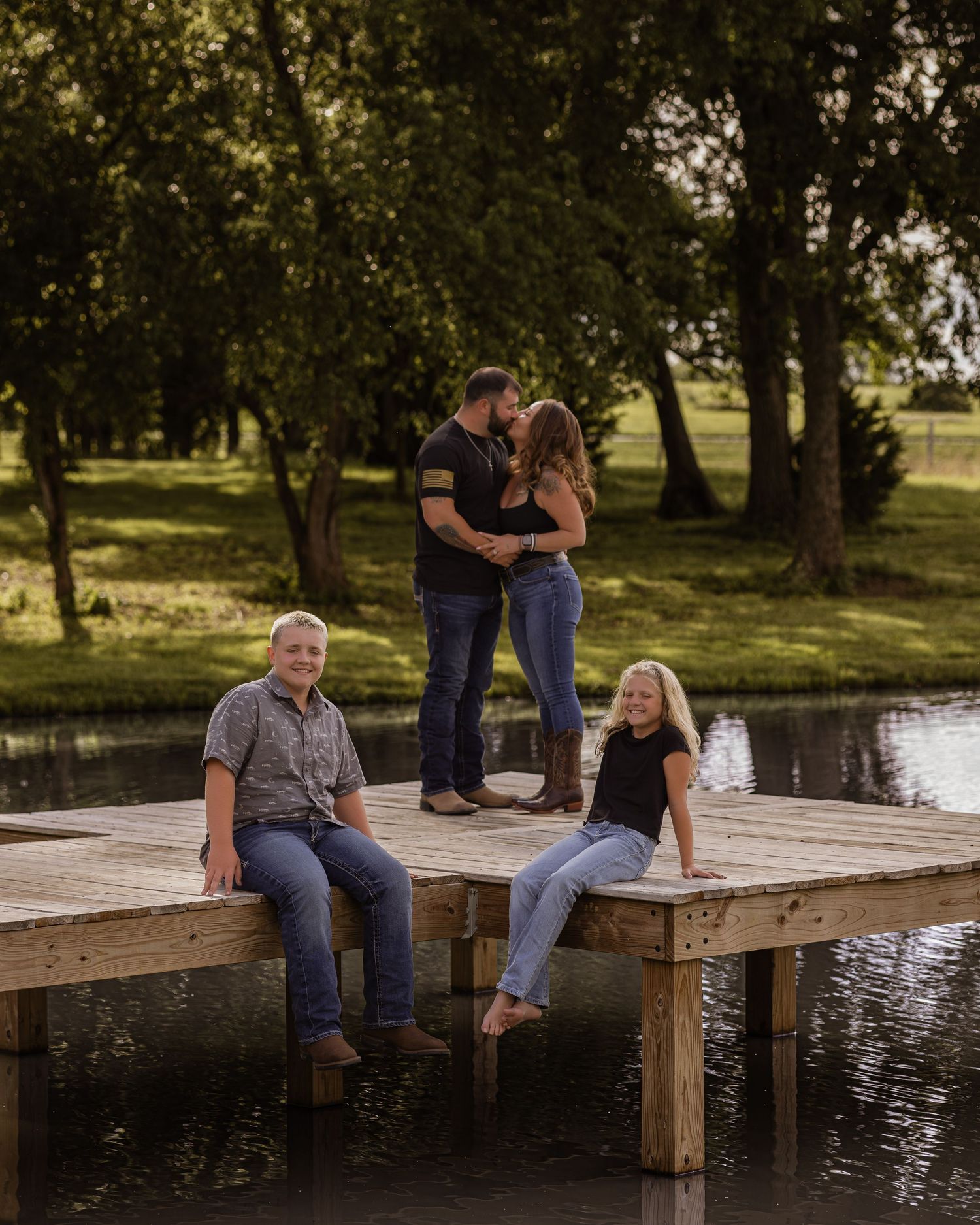 Family of four posing on a wooden dock by a pond, with parents kissing and two kids sitting on the edge.