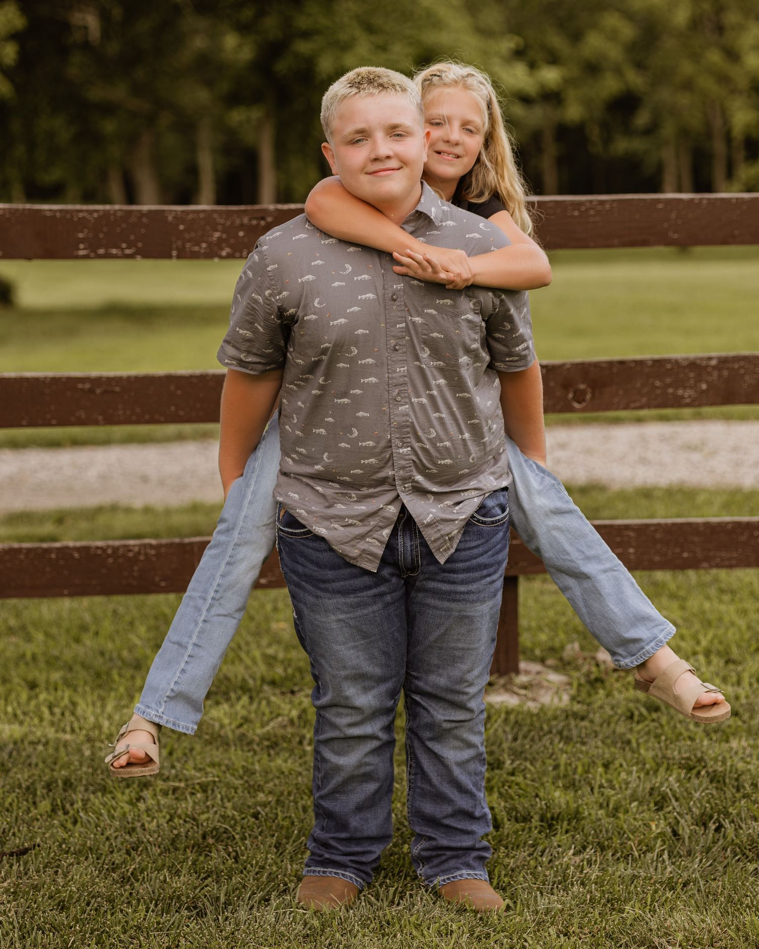 Boy giving a smiling girl a piggyback ride in front of a wooden fence on a green farm.