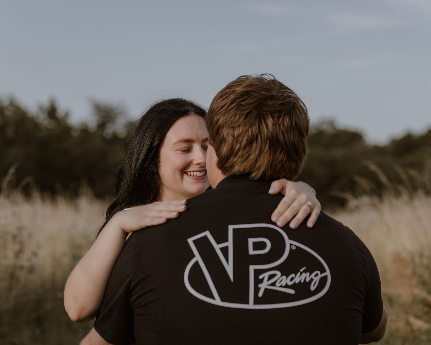 Happy woman hugging man wearing VP Racing shirt in golden field at dusk.