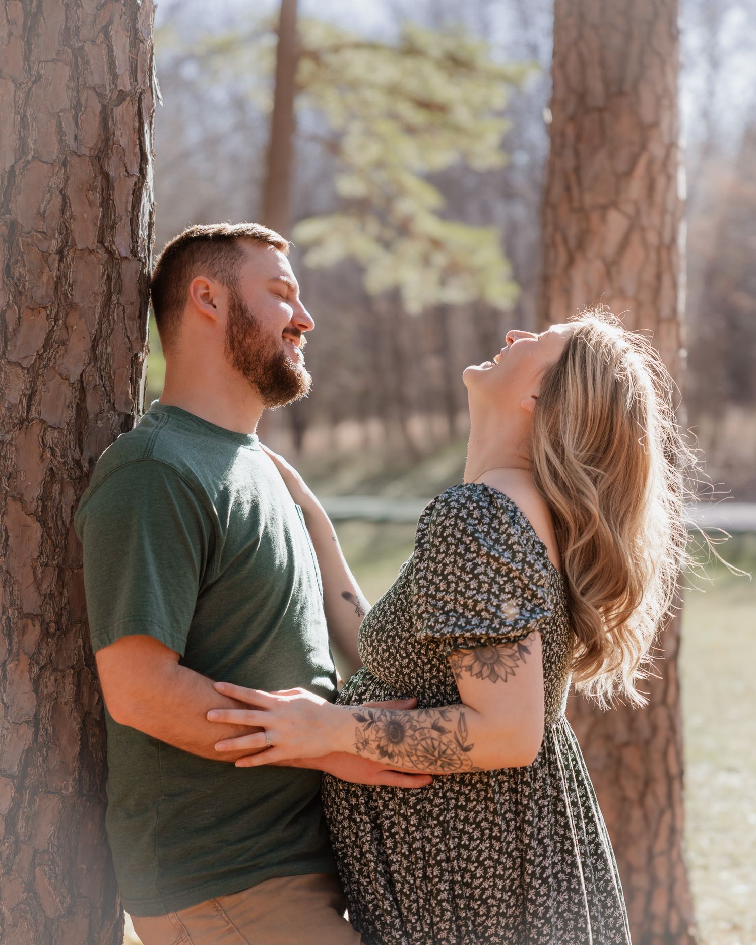 Joyful couple laughing together between tall pine trees in a sunlit forest setting.