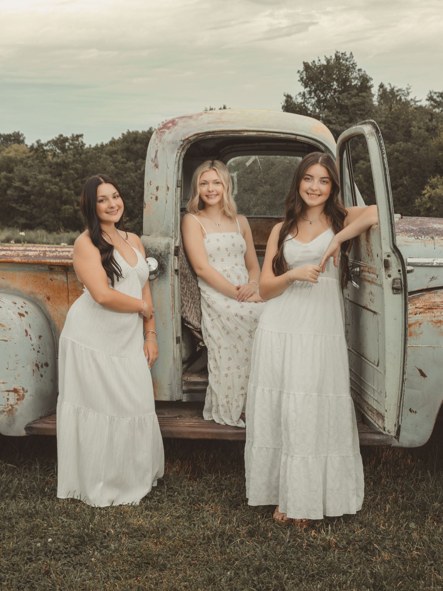 Three young women in white dresses pose by a vintage rusty truck in a scenic outdoor setting.