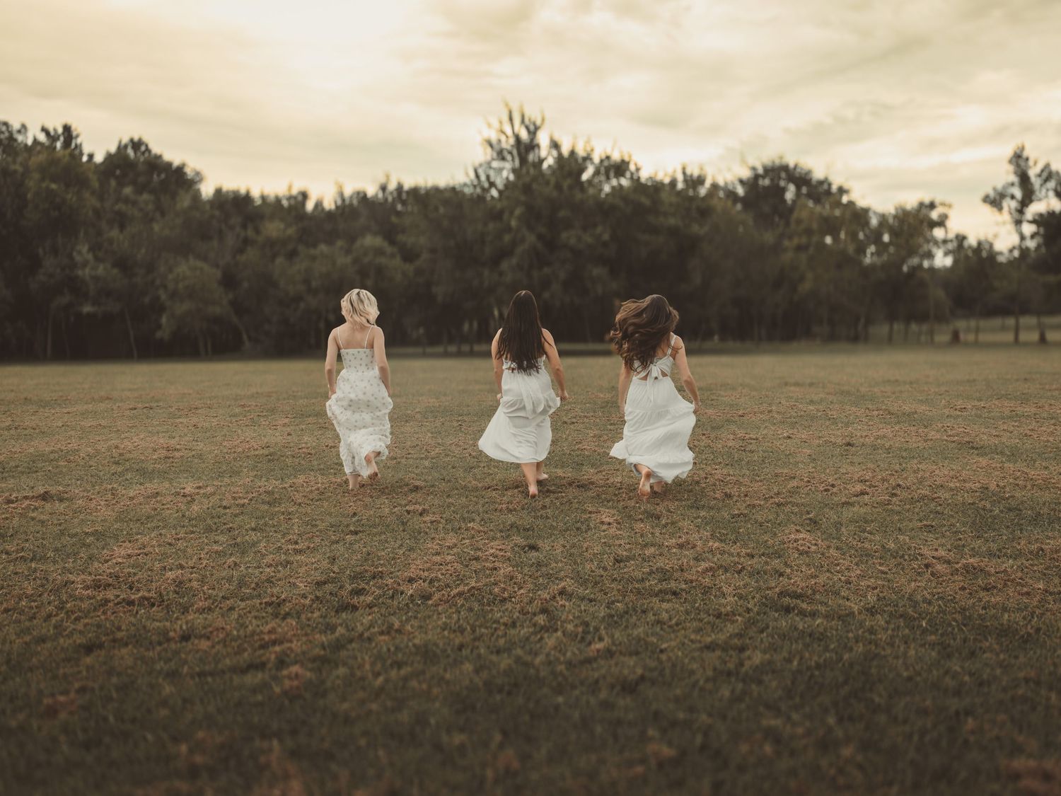 Three girls in white dresses running through a green field with trees in the background at dusk.