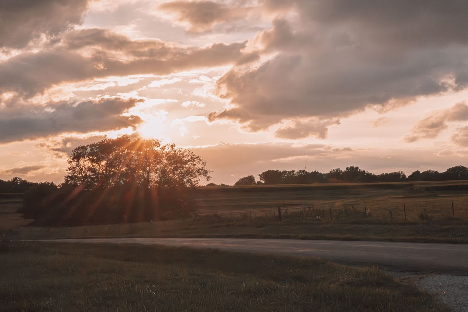Golden sunset rays burst through trees over a rural countryside road with dramatic cloudy sky.