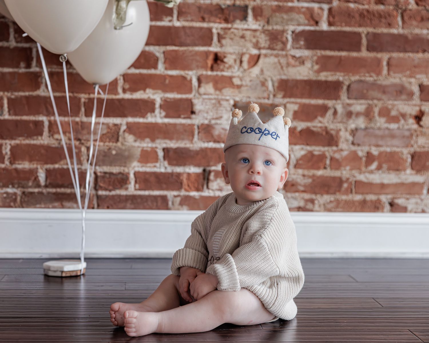 Baby boy named Cooper sits against brick wall wearing a personalized crown hat with white balloons for his first birthday.
