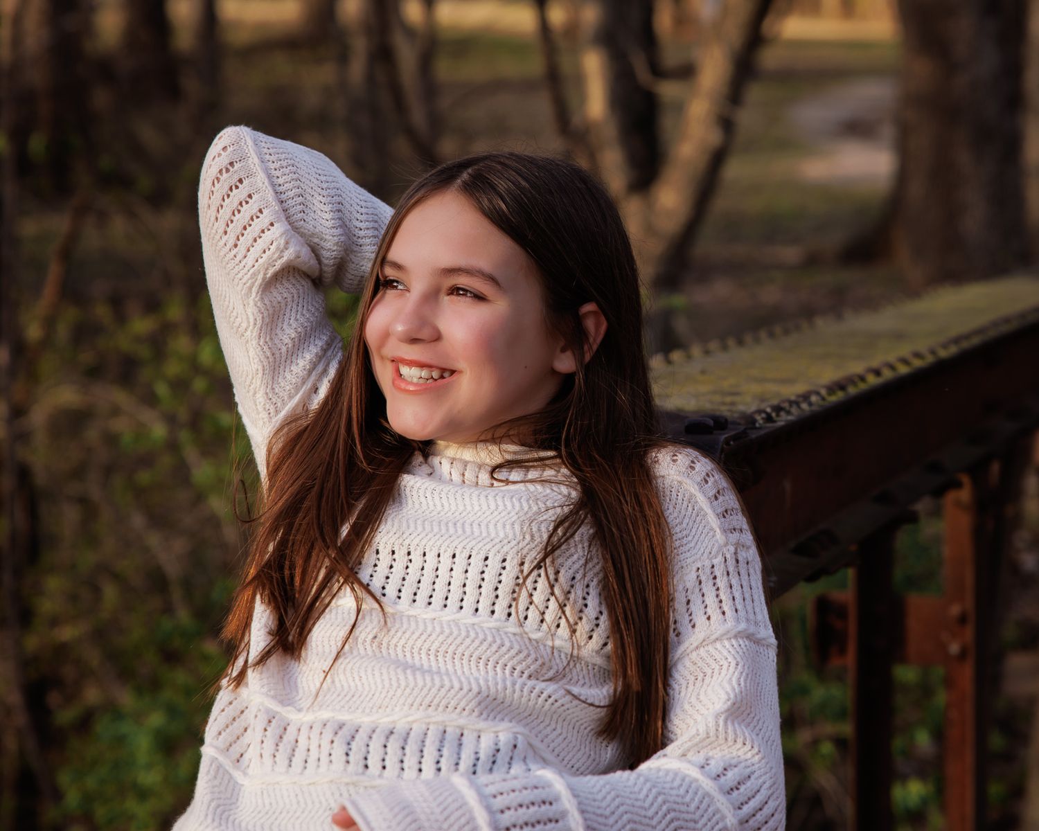 Woman in white knit sweater relaxes outdoors while sitting on a wooden bench in a park setting.