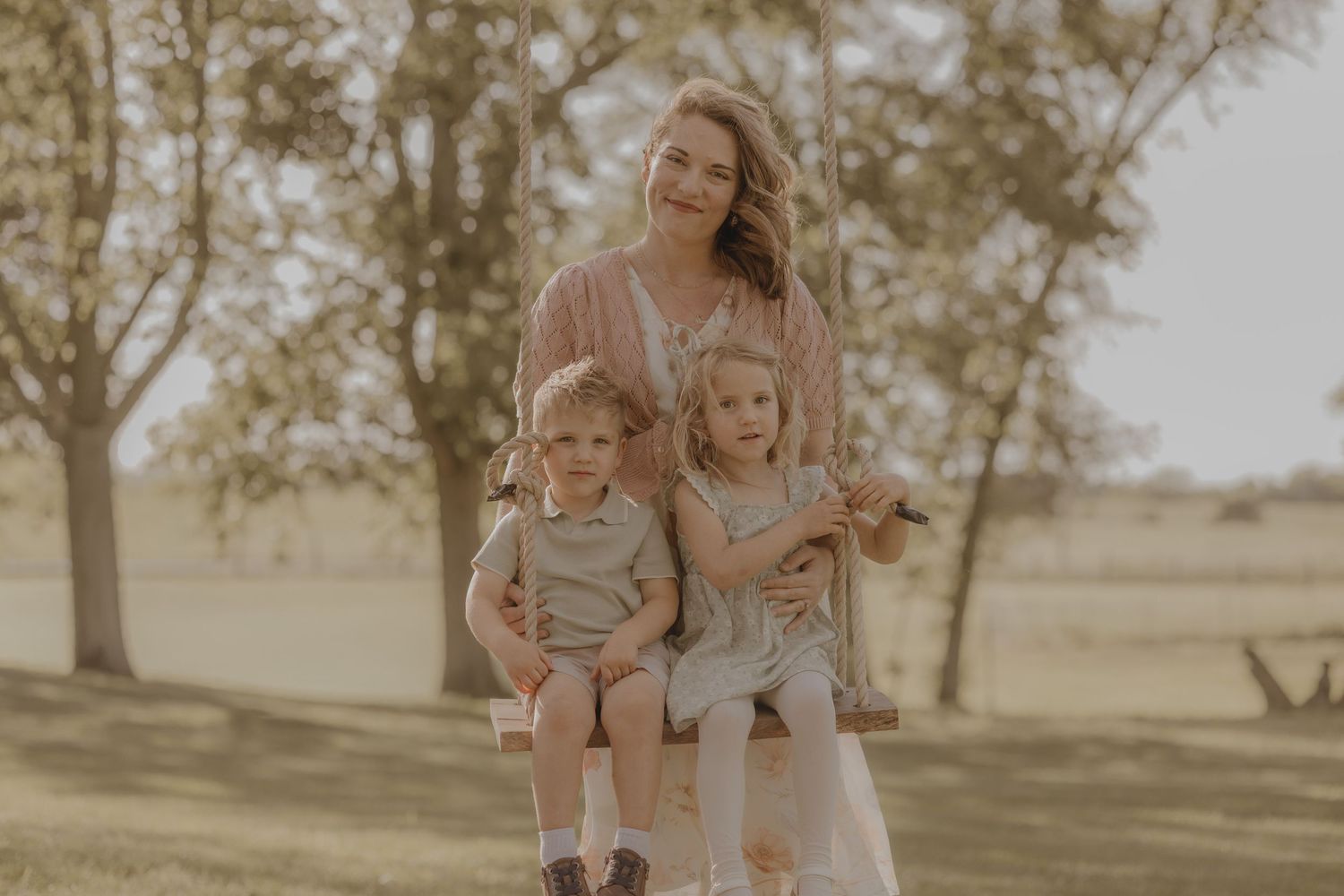A smiling mother sits on a swing with her two young children in a sunny outdoor setting.