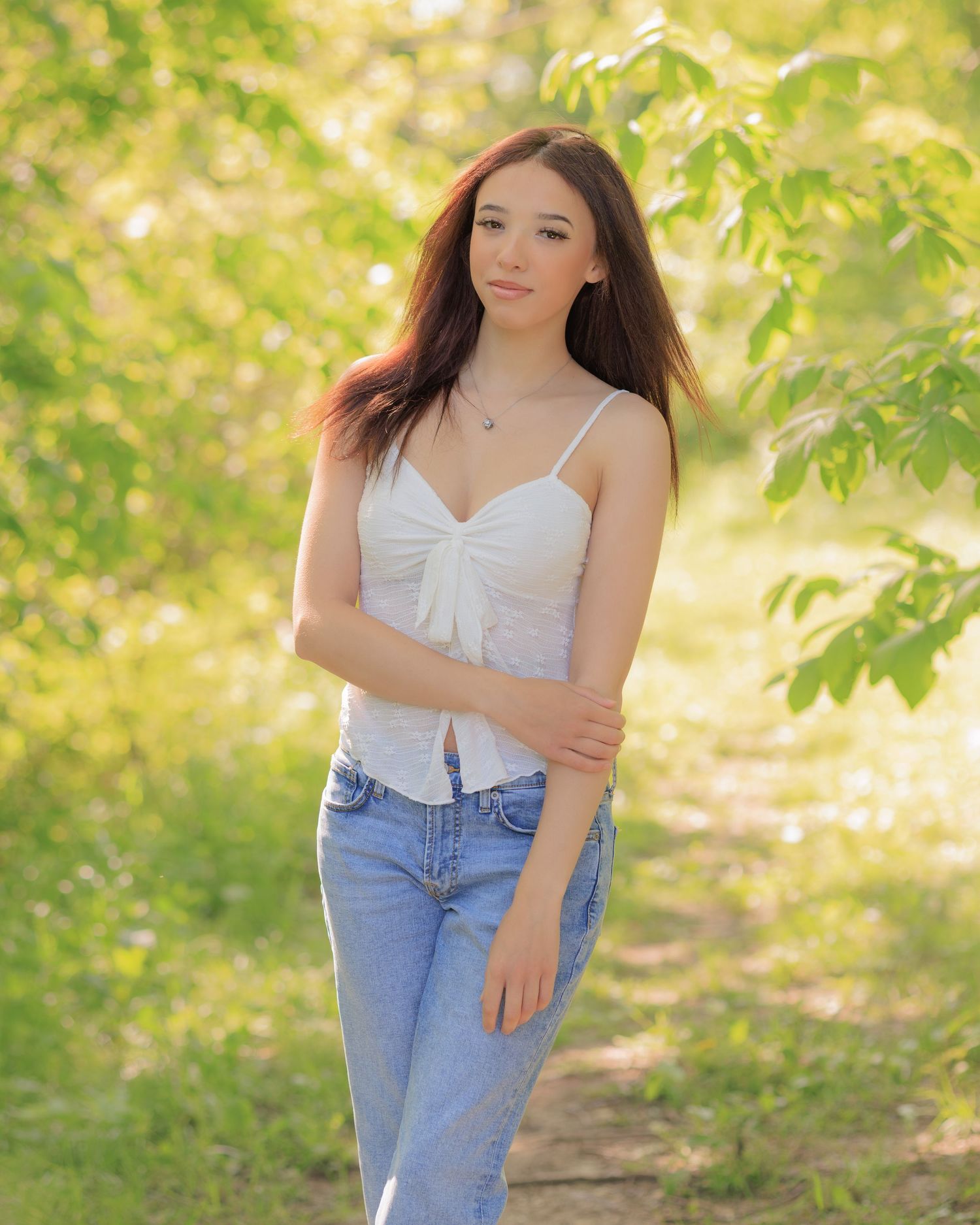 Young woman in white top and jeans poses outdoors among lush green trees on a sunny day.