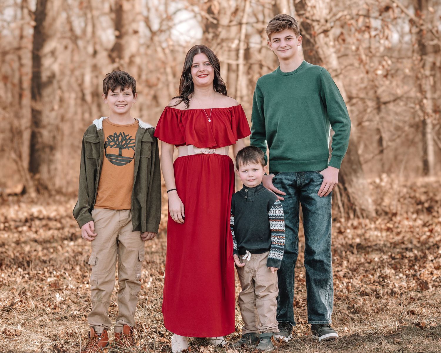 A small family group poses outdoors in a wooded setting wearing coordinated casual clothing with fall foliage behind them.