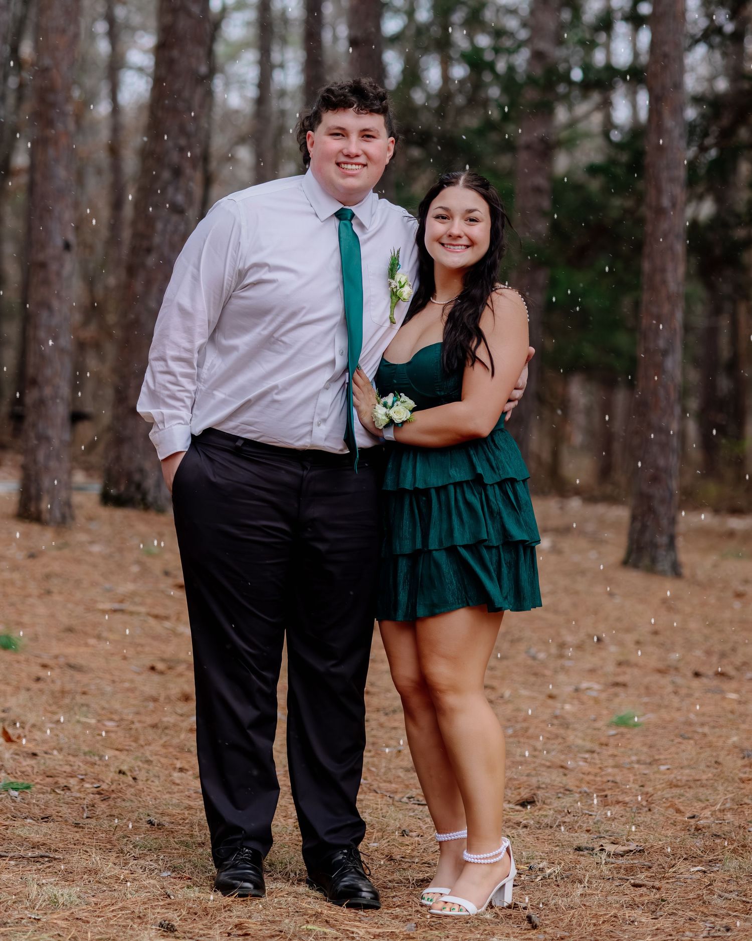 A couple in formal attire poses playfully in a forest setting, wearing emerald green and white outfits.
