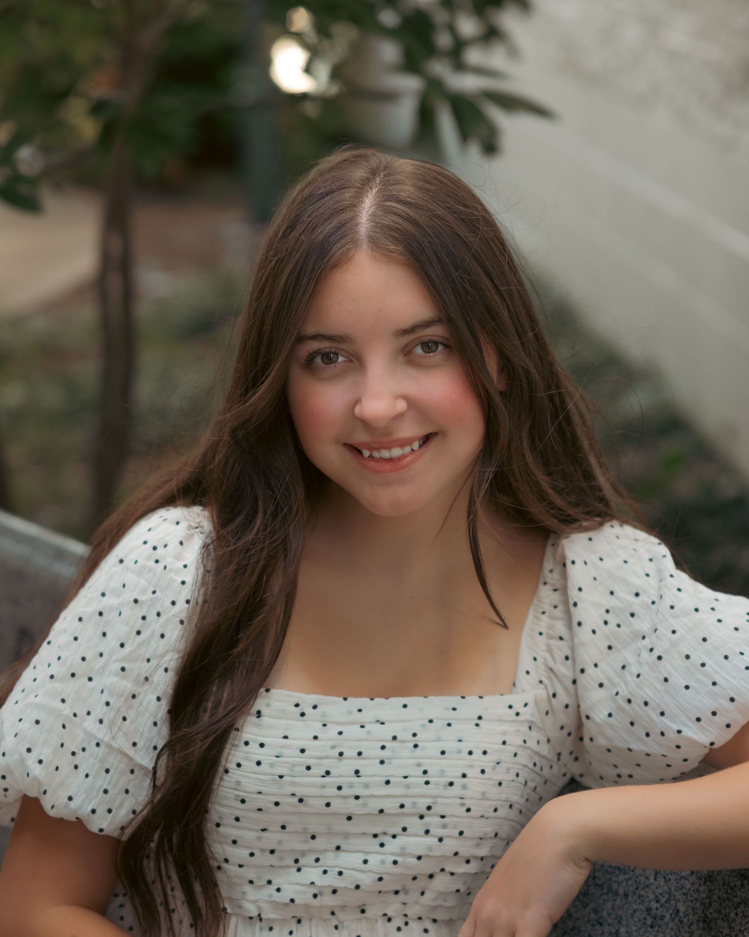 Young woman with long brown hair smiling outdoors, wearing a white polka dot dress with puff sleeves.