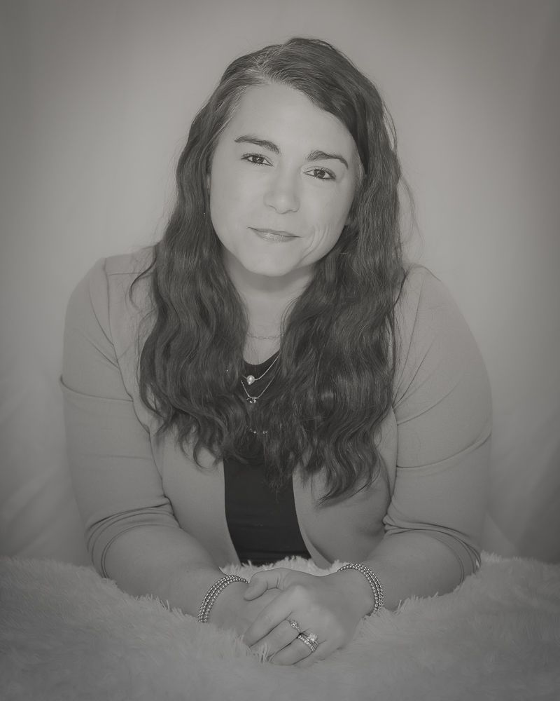 Black and white portrait of a young person with long dark wavy hair wearing a casual top and sitting with hands clasped.