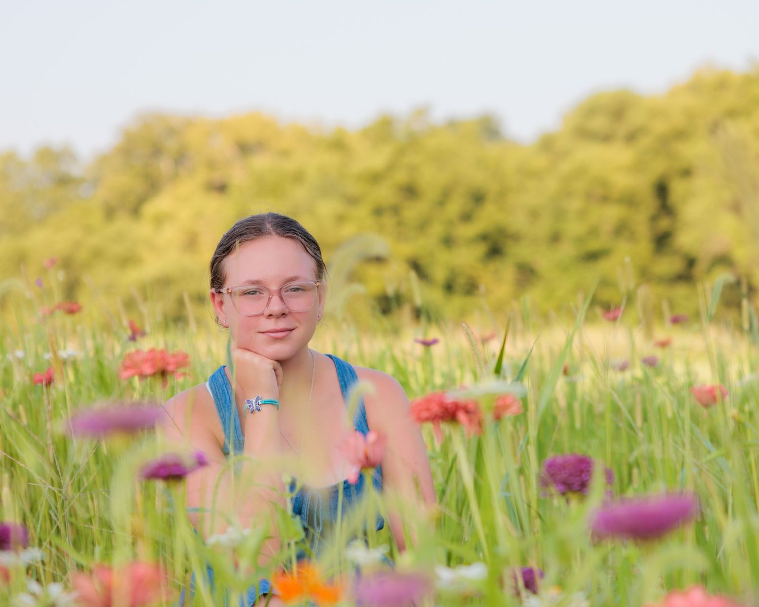 Young girl with glasses sitting in a vibrant wildflower meadow with colorful blooms and lush green trees in background.
