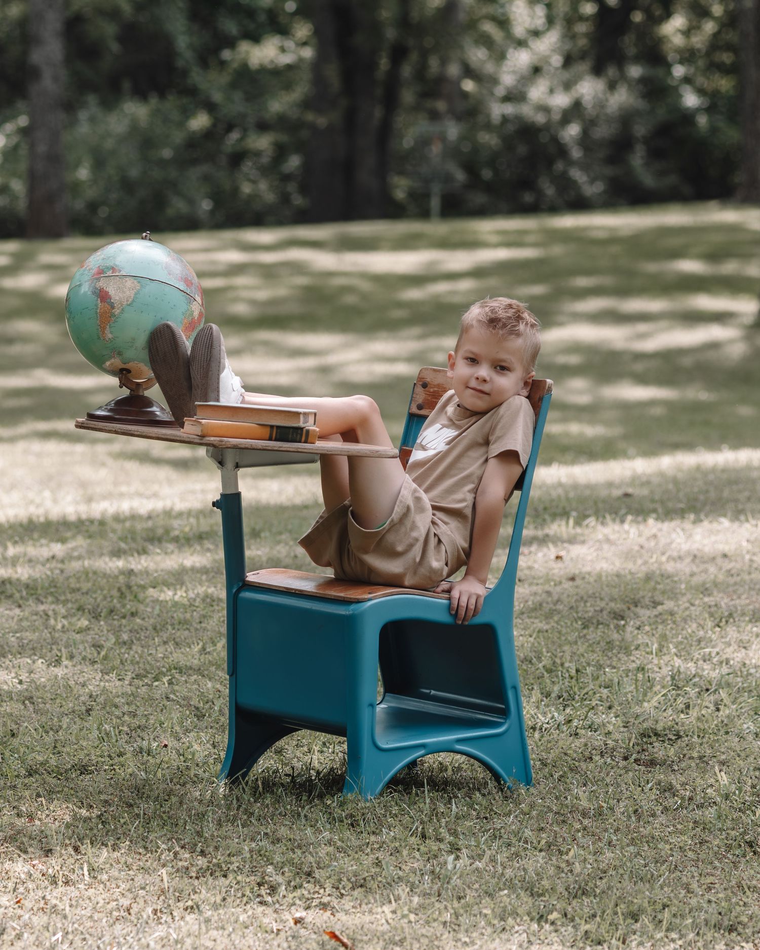 Young boy sitting in a teal school desk outdoors with a vintage globe, wearing beige outfit in a grassy park setting.