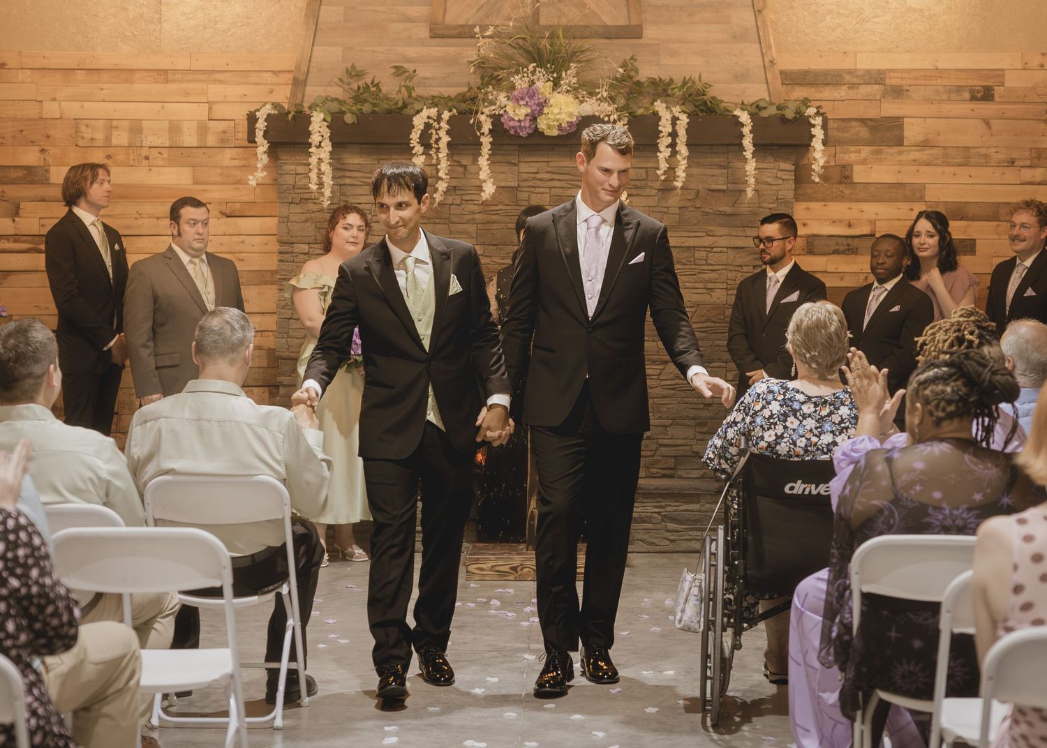 Two grooms walk hand-in-hand down the aisle at their wedding ceremony, surrounded by applauding guests.