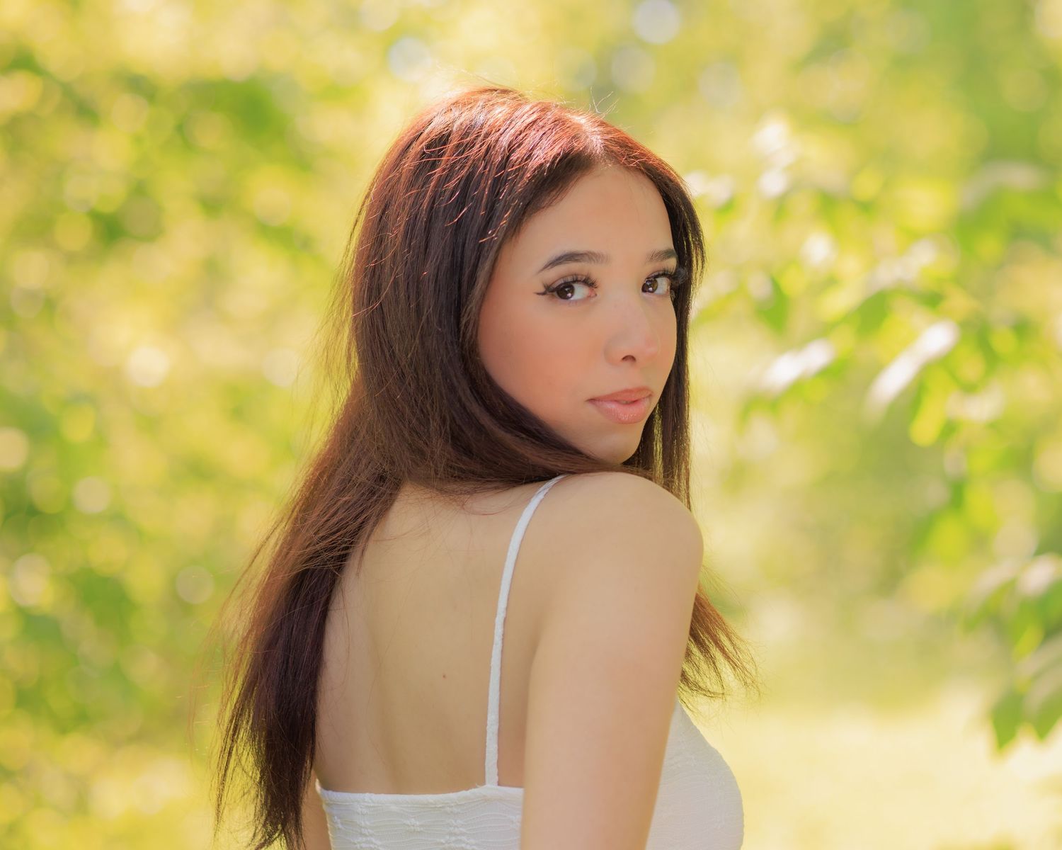 Young woman with long brown hair in white dress poses outdoors among lush green trees in soft natural light.