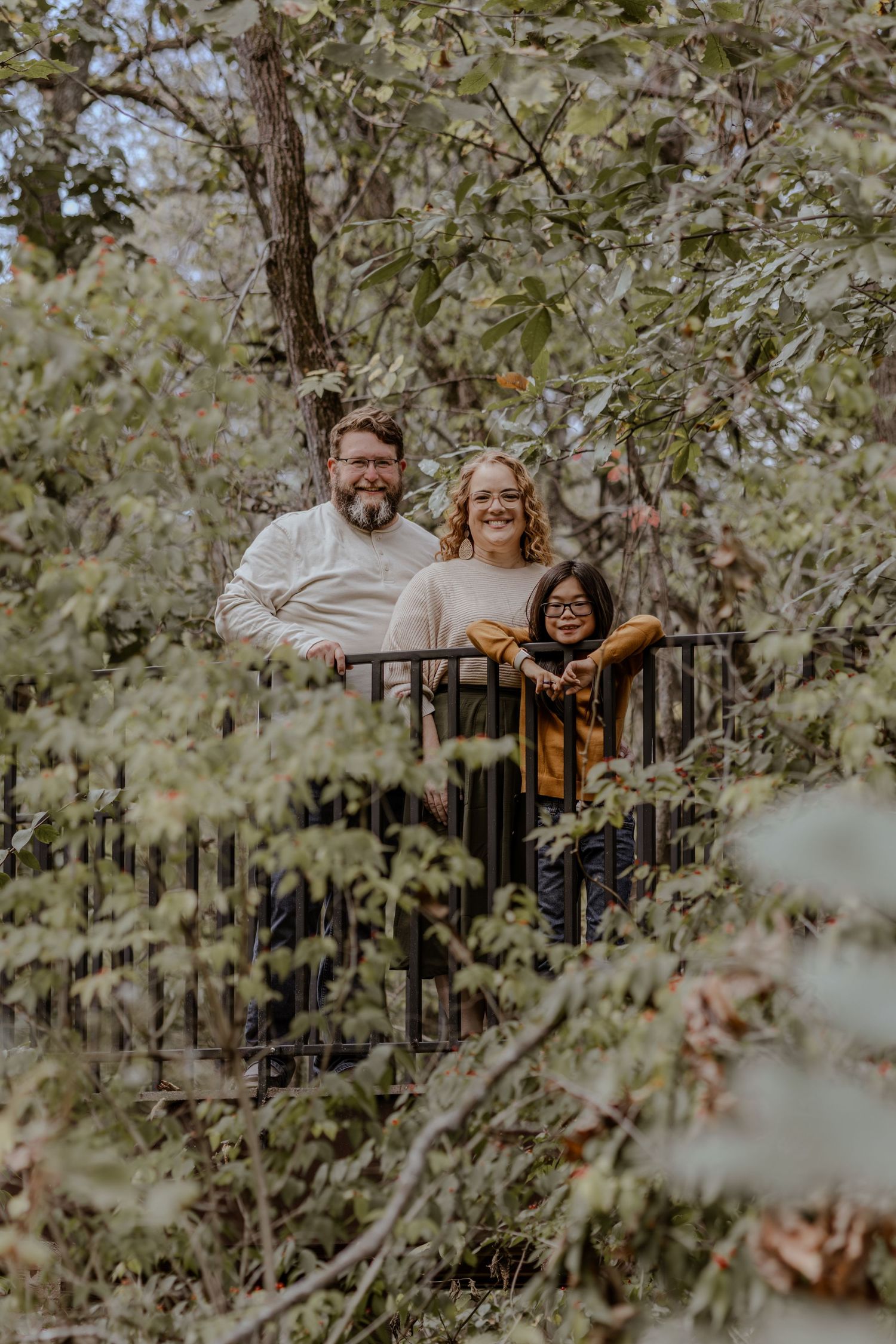 Family of three smiling behind a black railing surrounded by lush green trees in an outdoor setting.