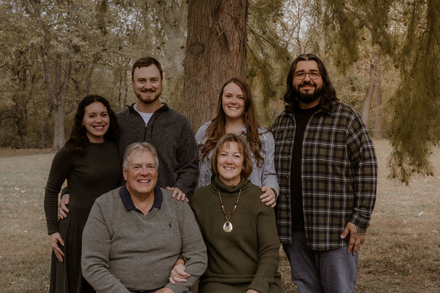 A family group poses together outdoors in a wooded setting on an autumn day.