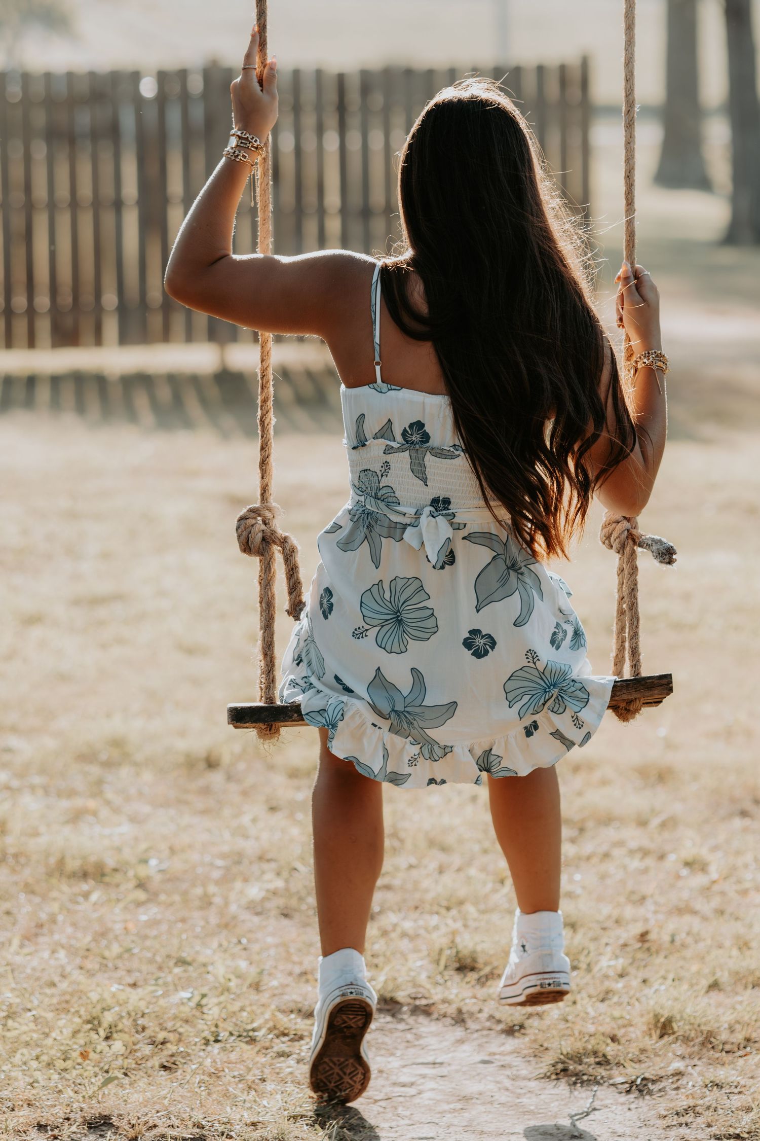 Young girl in floral romper swinging on a rustic rope swing outdoors in a sun-drenched playground.