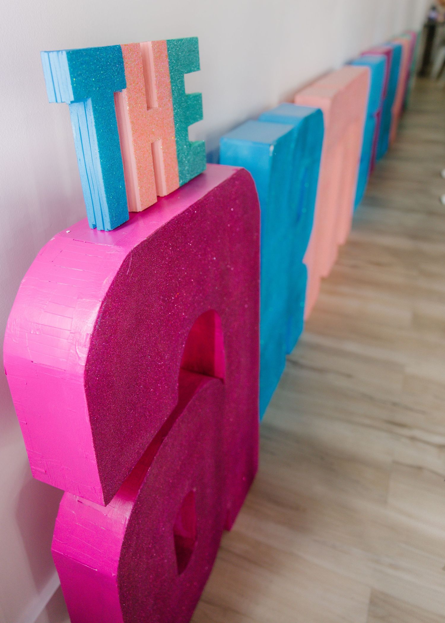 Large colorful foam letters in pink, blue, and orange spelling a phrase, displayed on a light wood floor.