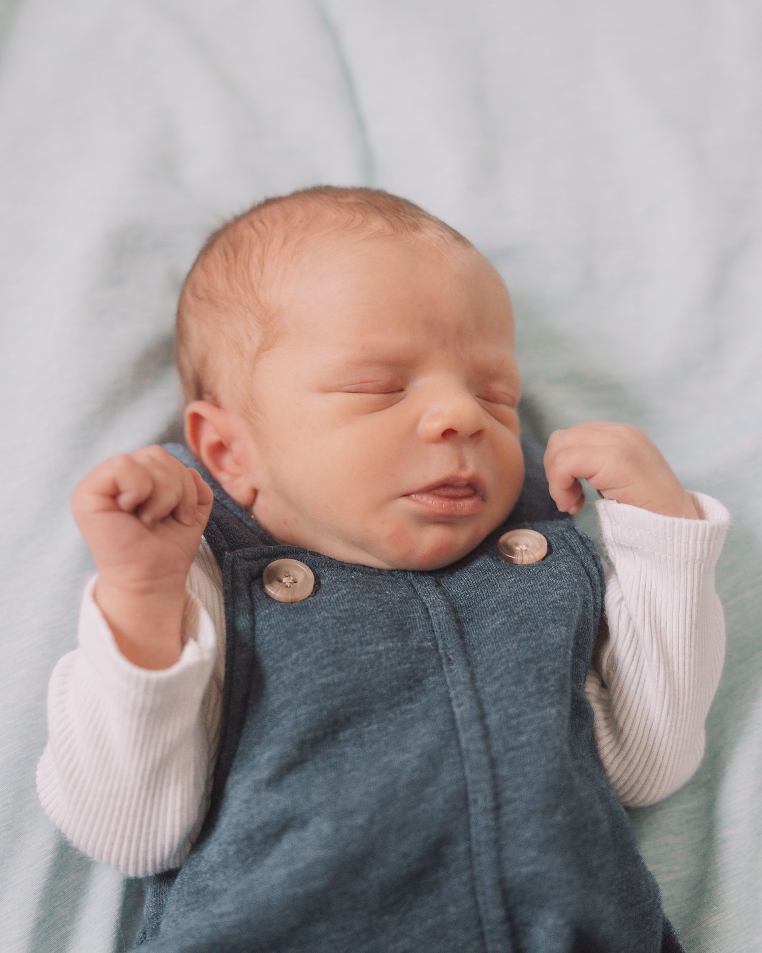 Peaceful newborn baby sleeping in a teal vest with wooden buttons over a white long-sleeve onesie.