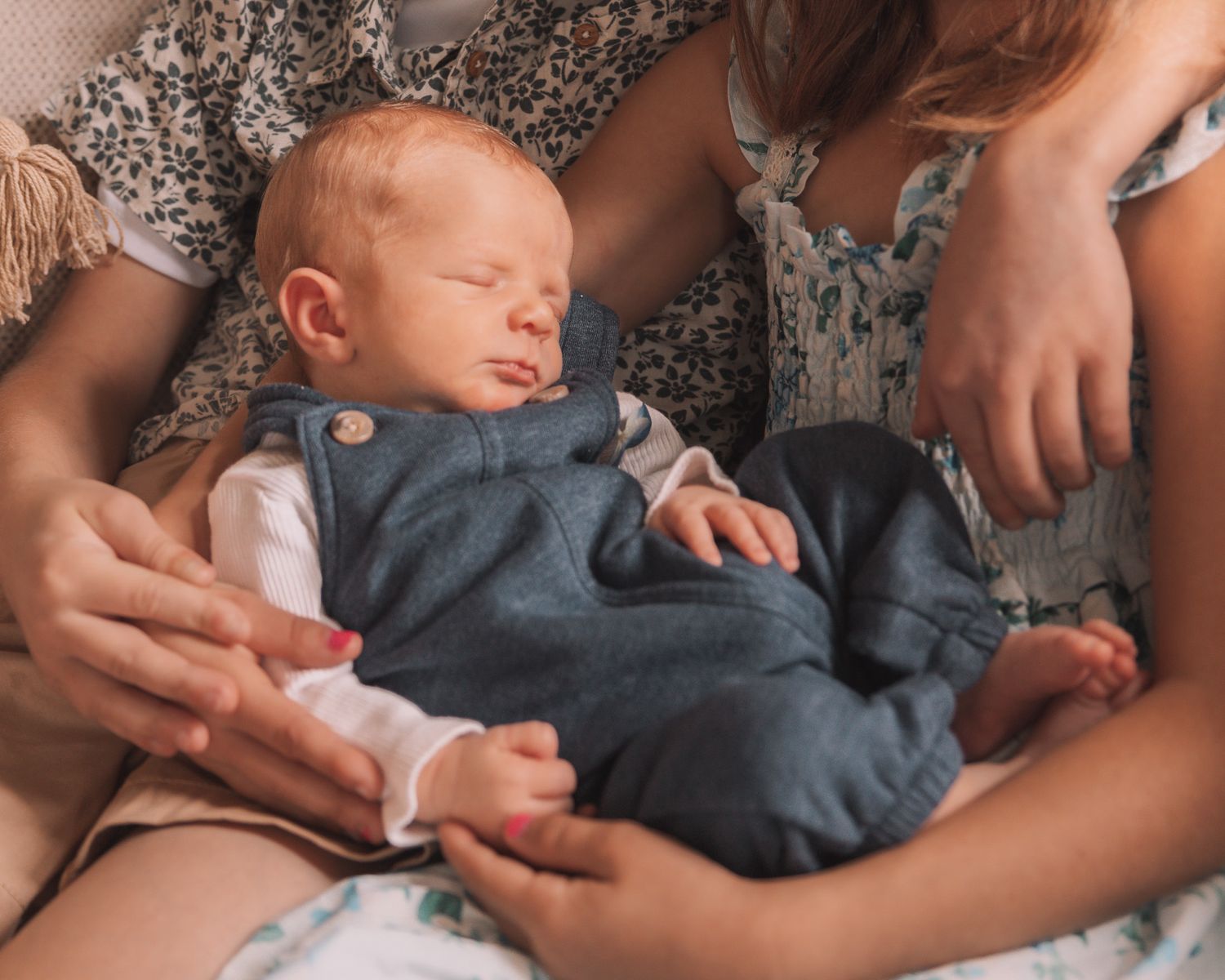 Two adults gently holding a sleeping redhead newborn baby in a blue denim romper.