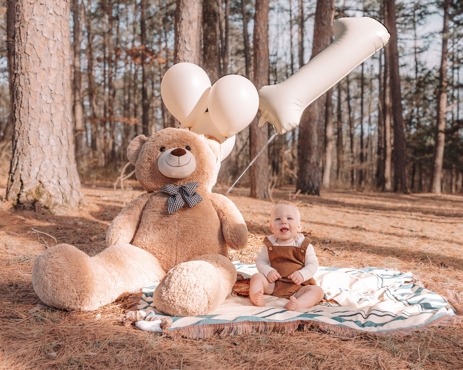 A large teddy bear and smaller stuffed animal sit on a blanket in a pine forest setting.