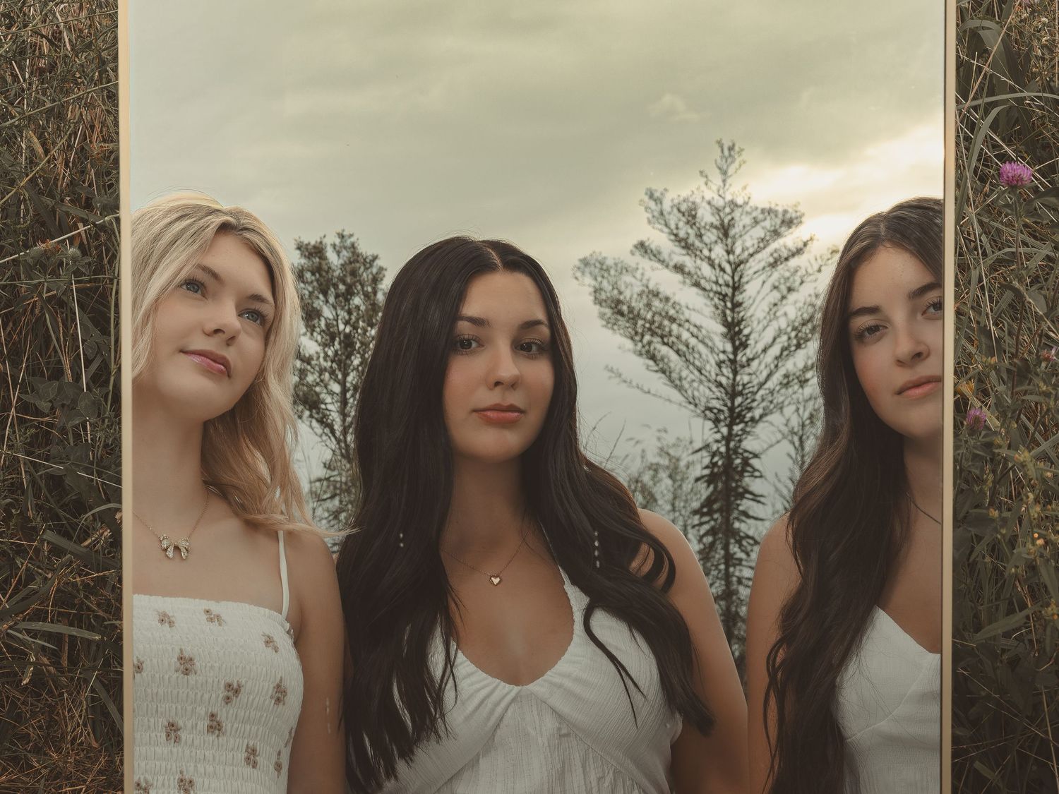 Three young women in white dresses pose outdoors surrounded by tall grass and trees under a cloudy sky.