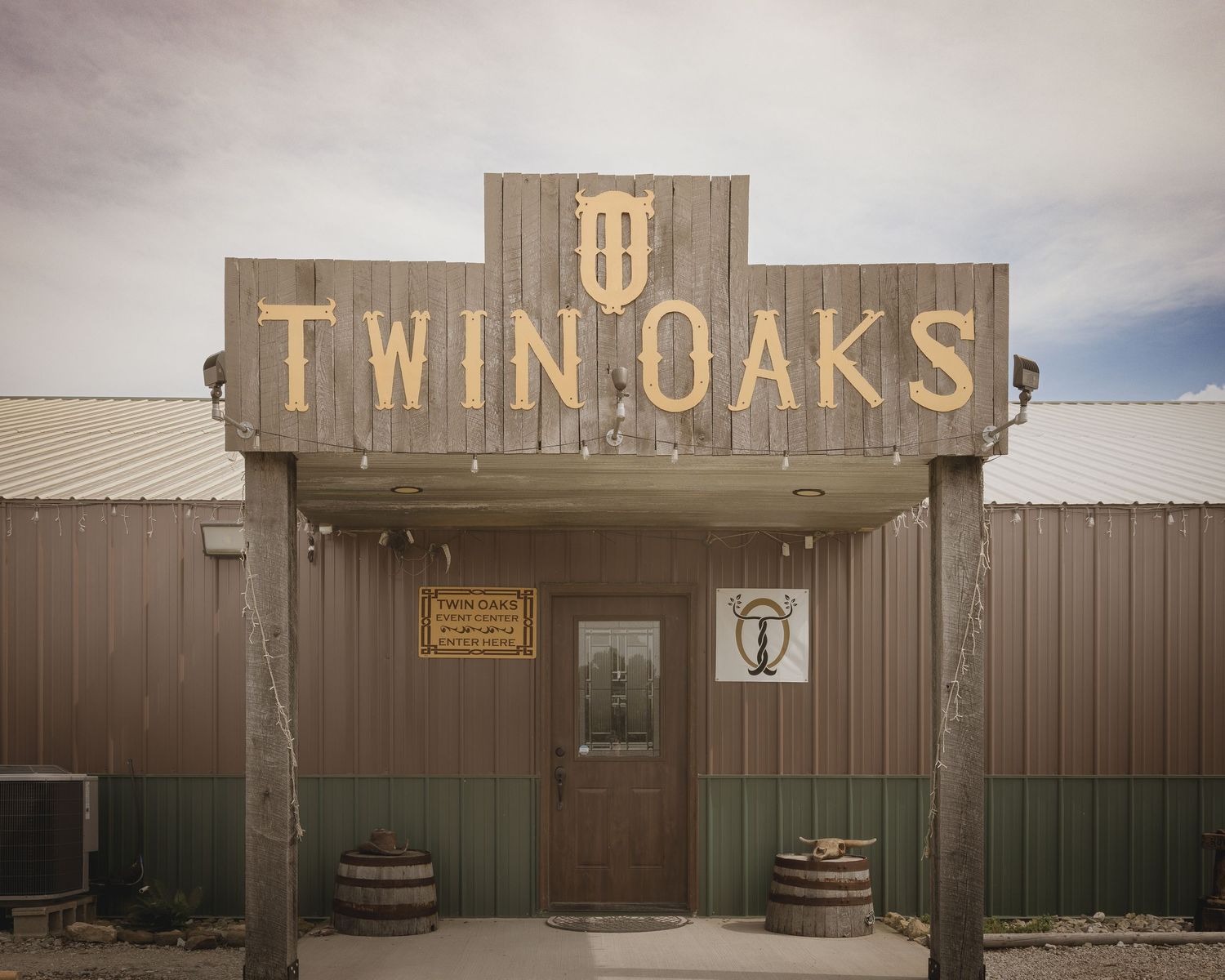 Rustic Twin Oaks venue exterior with wooden sign, metal siding, and barrel decorations under cloudy sky.