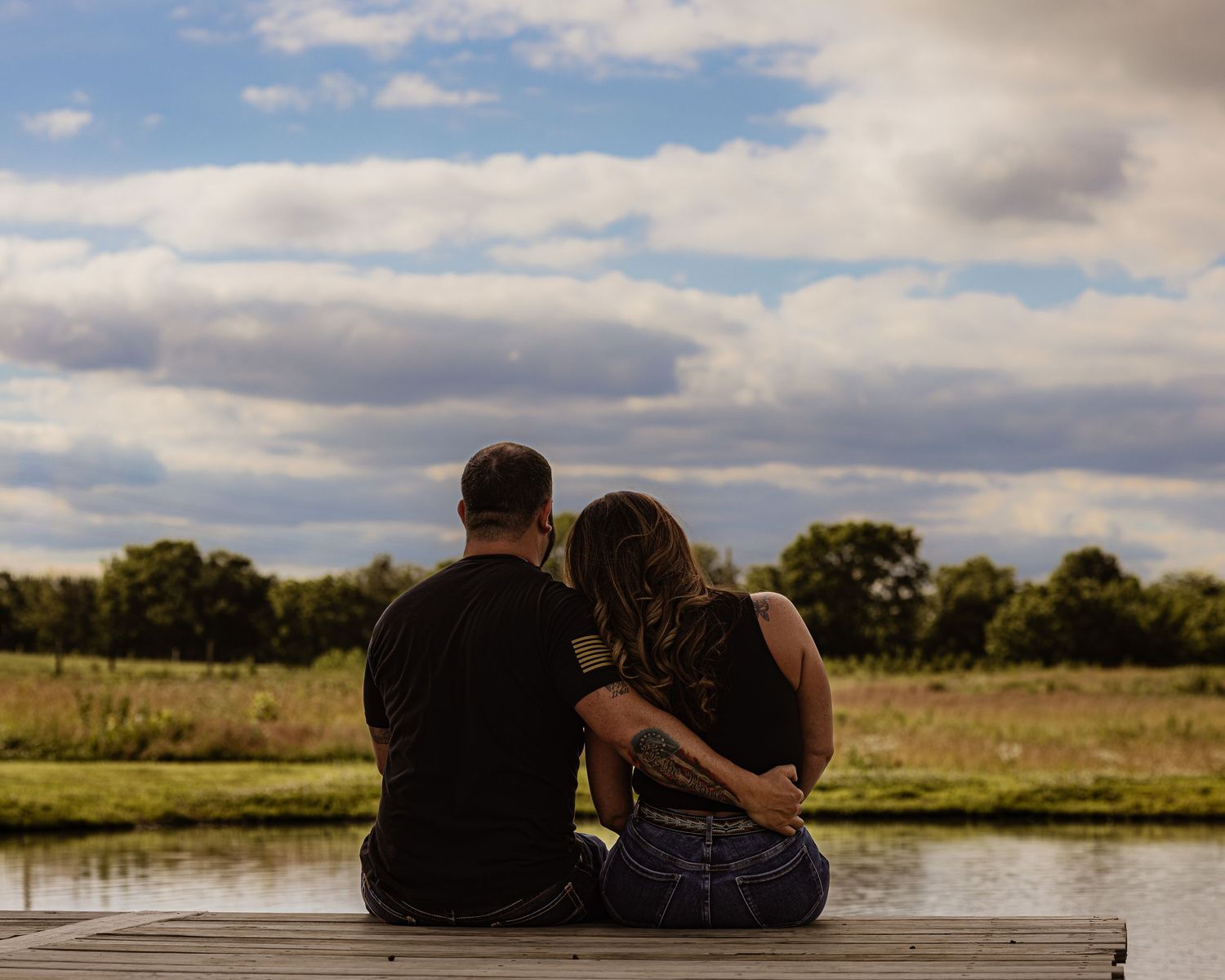 Couple sitting together on a dock overlooking a scenic pond under a dramatic cloudy sky.