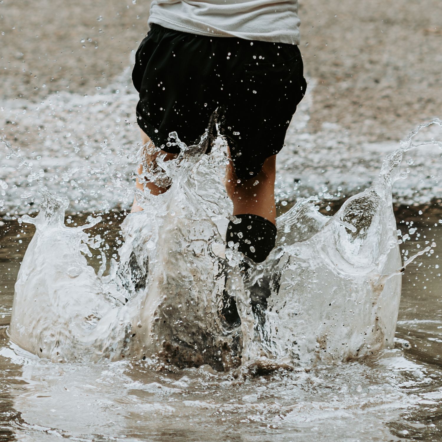 Child splashing through shallow water puddle on sandy beach, wearing black shorts and boots, creating dynamic water spray.