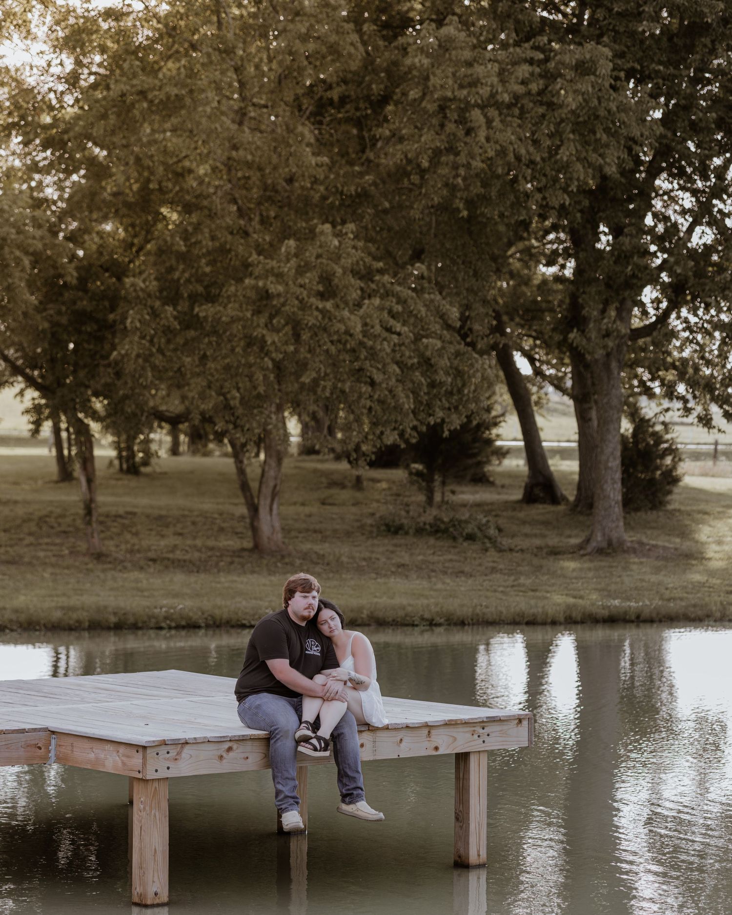 A couple sits closely together on a rustic wooden dock beside a serene pond with tall trees in background.