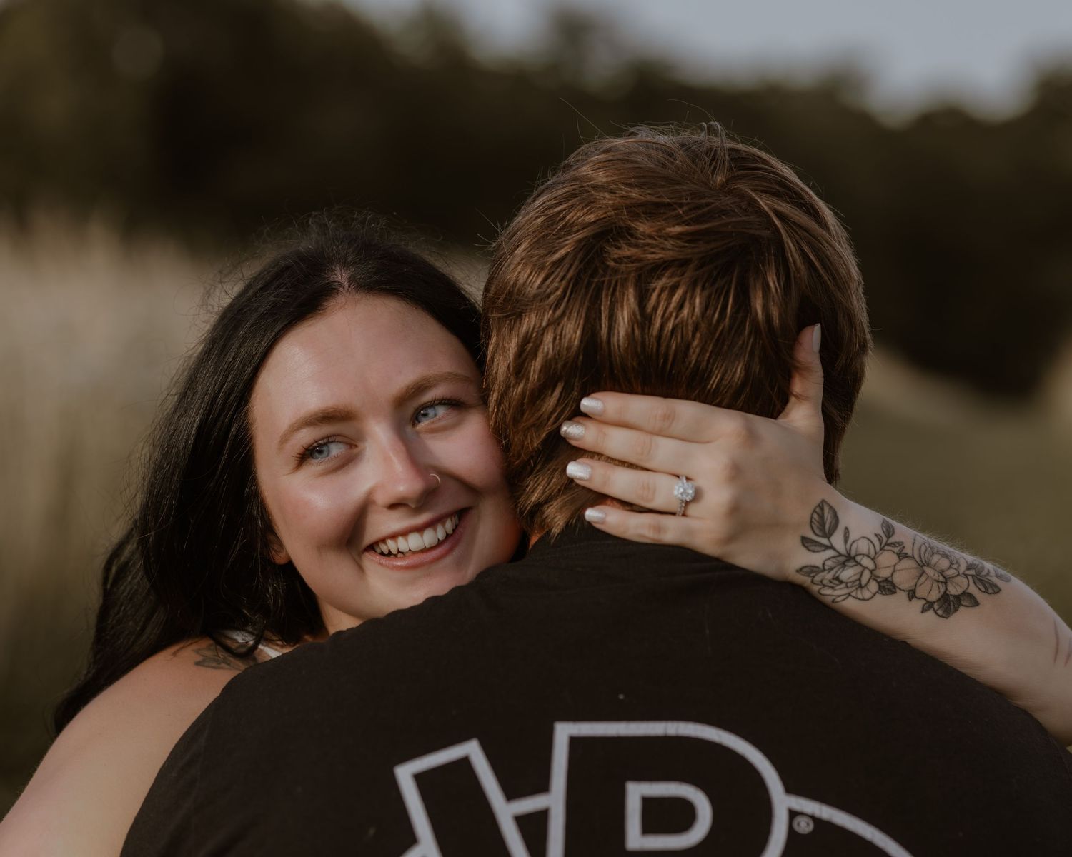 Happy woman hugging her partner in a sunlit field, showing engagement ring.