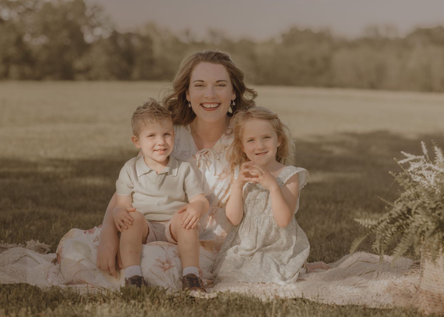 A mother smiles warmly with her two young children sitting on a blanket in a sunny green field.
