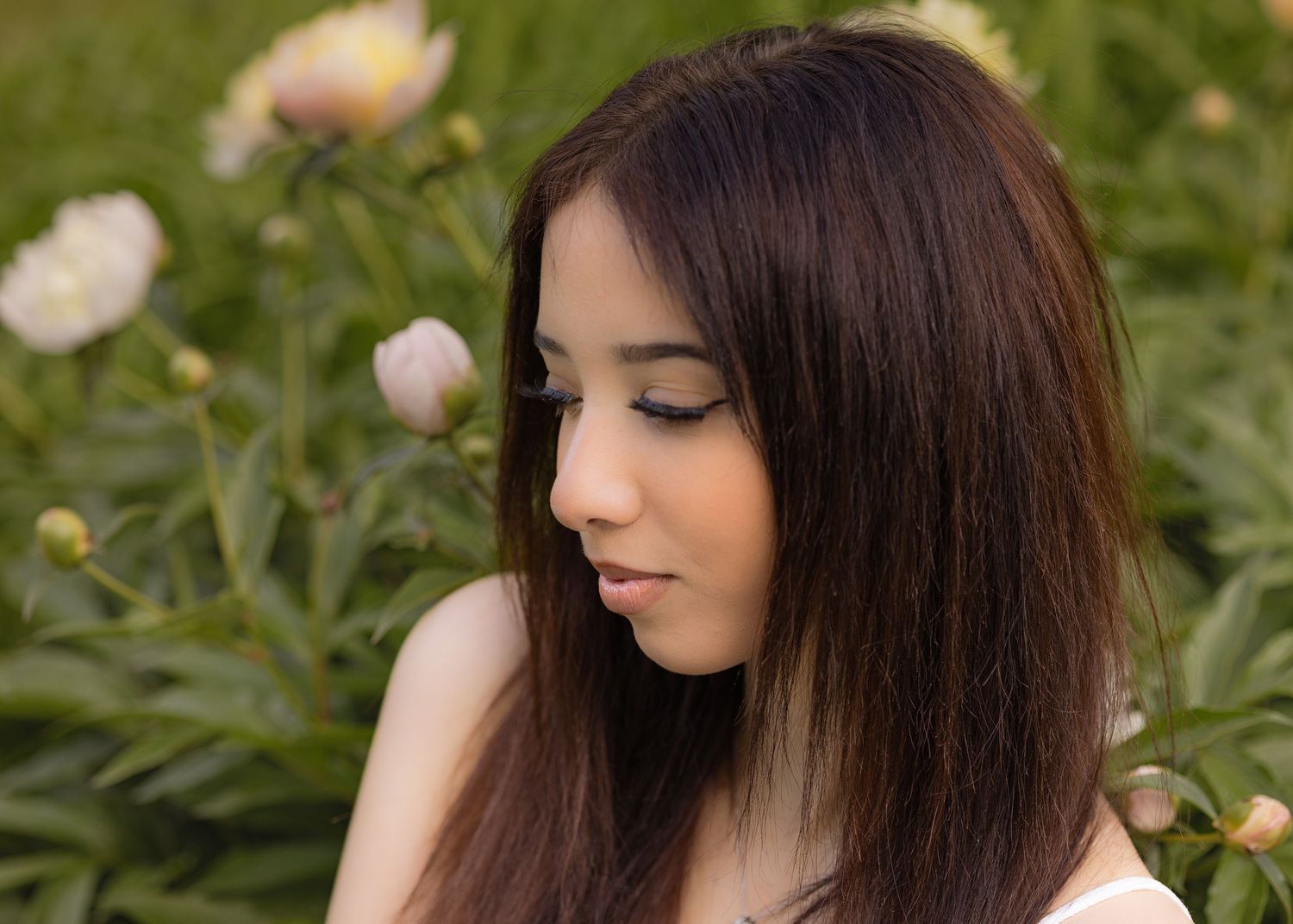 Young woman with long brown hair gazing downward surrounded by blooming pink peonies in a lush garden.