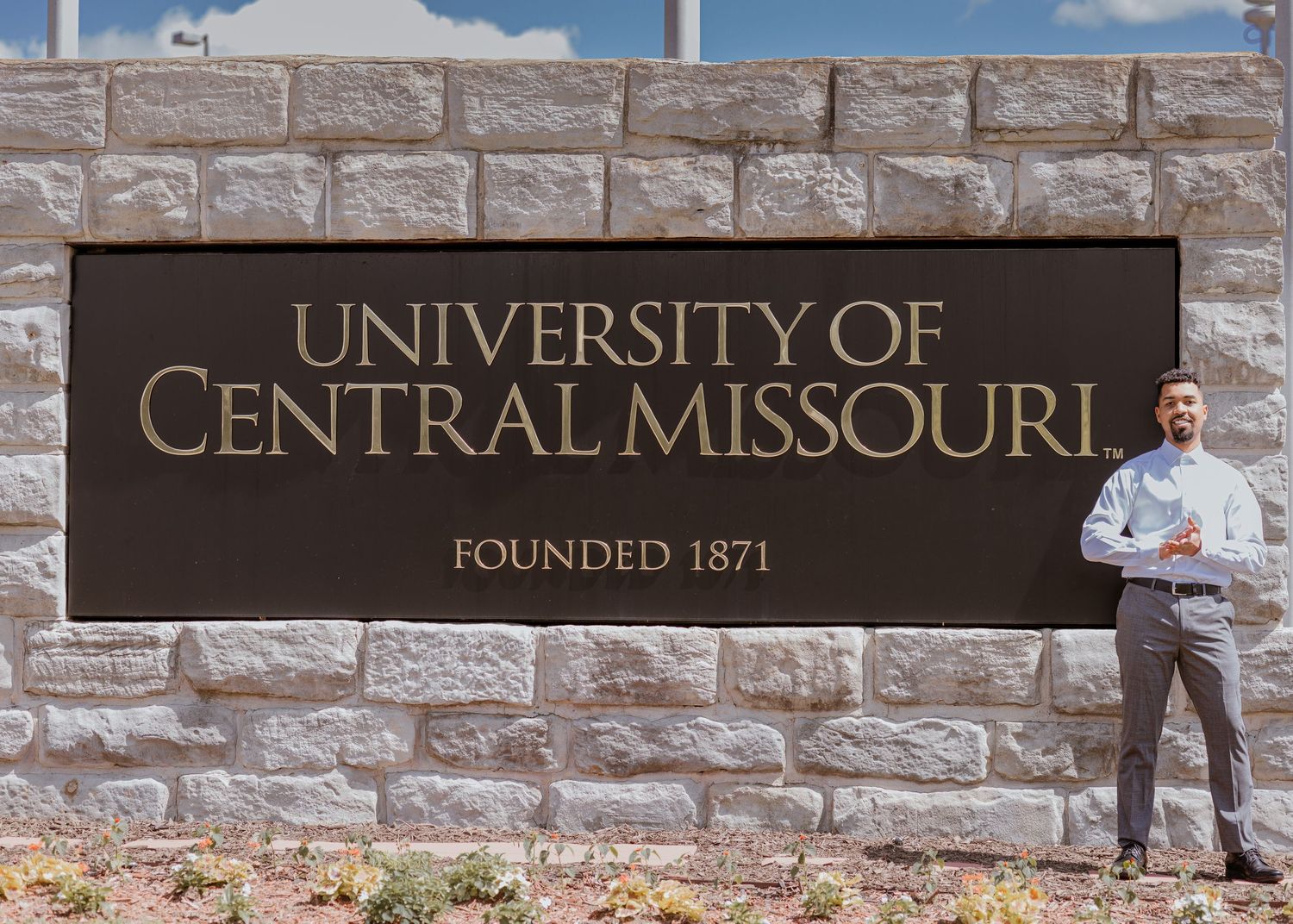 Man standing beside University of Central Missouri founded 1871 stone entrance sign on sunny day.