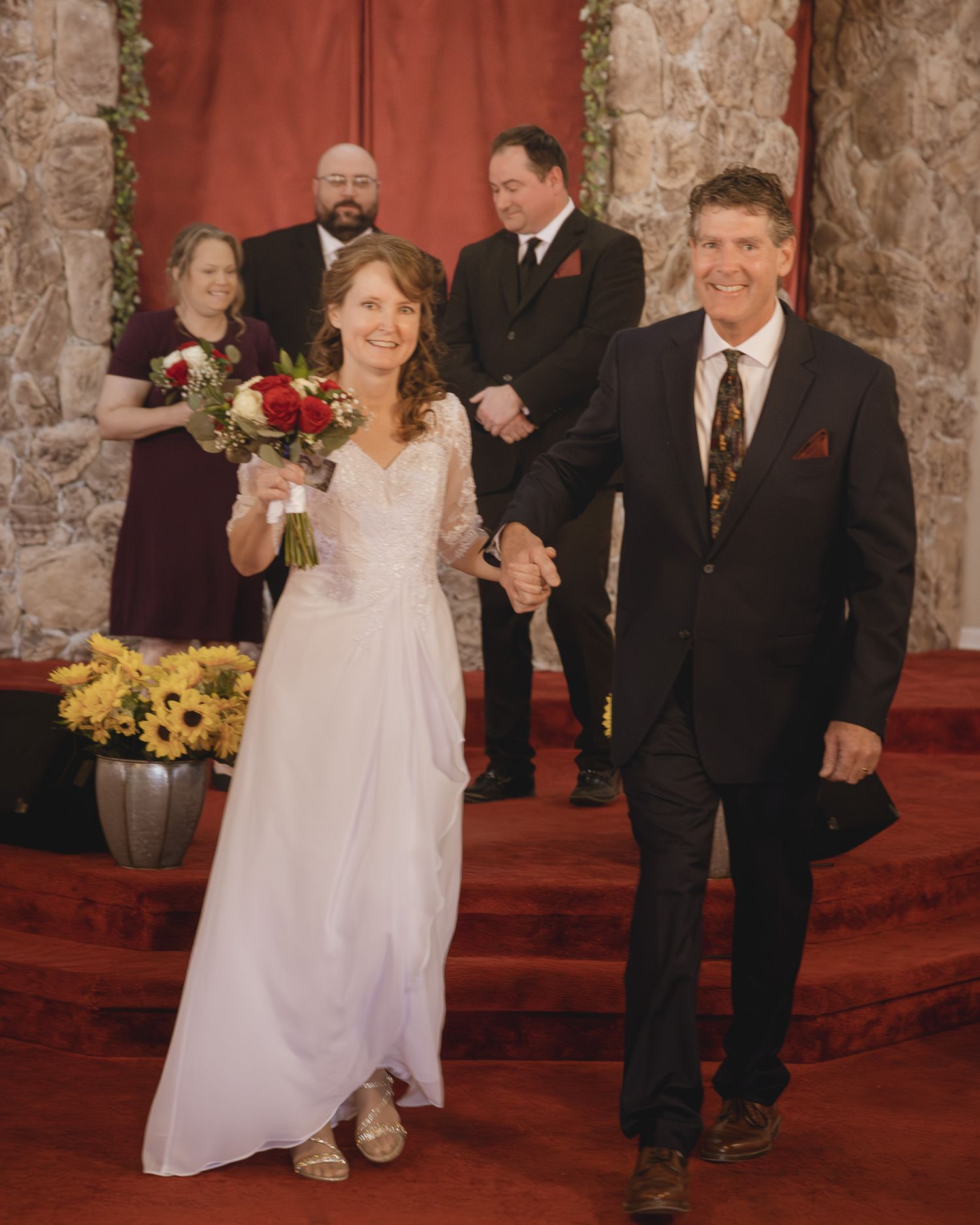 Wedding ceremony procession on red carpeted church steps with stone walls and floral decorations.