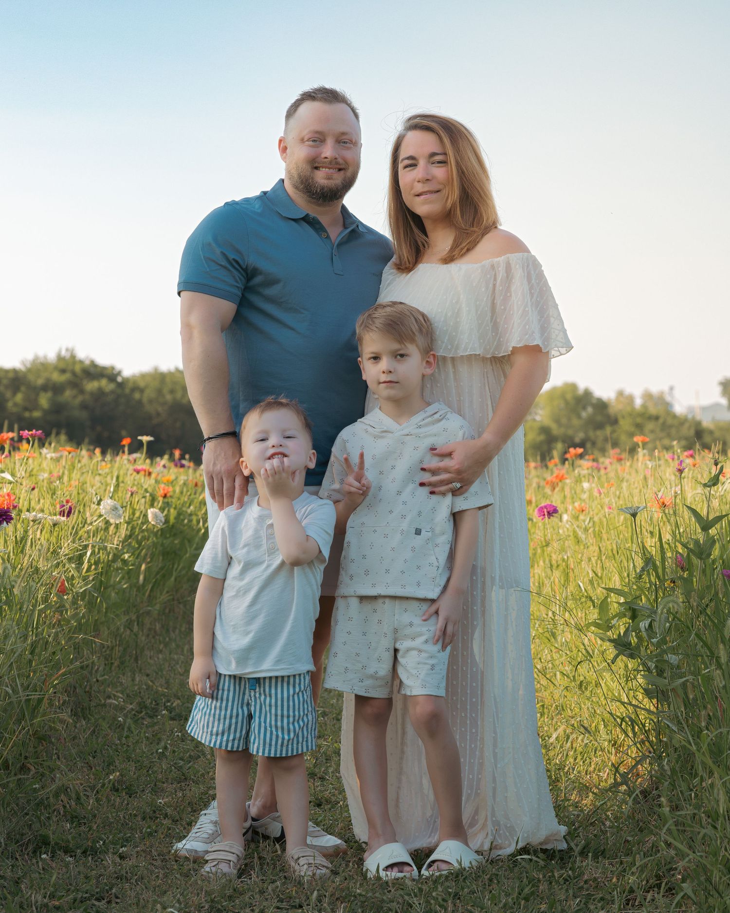 Family of four poses in a sunny wildflower field, parents with two young boys wearing coordinated outfits.
